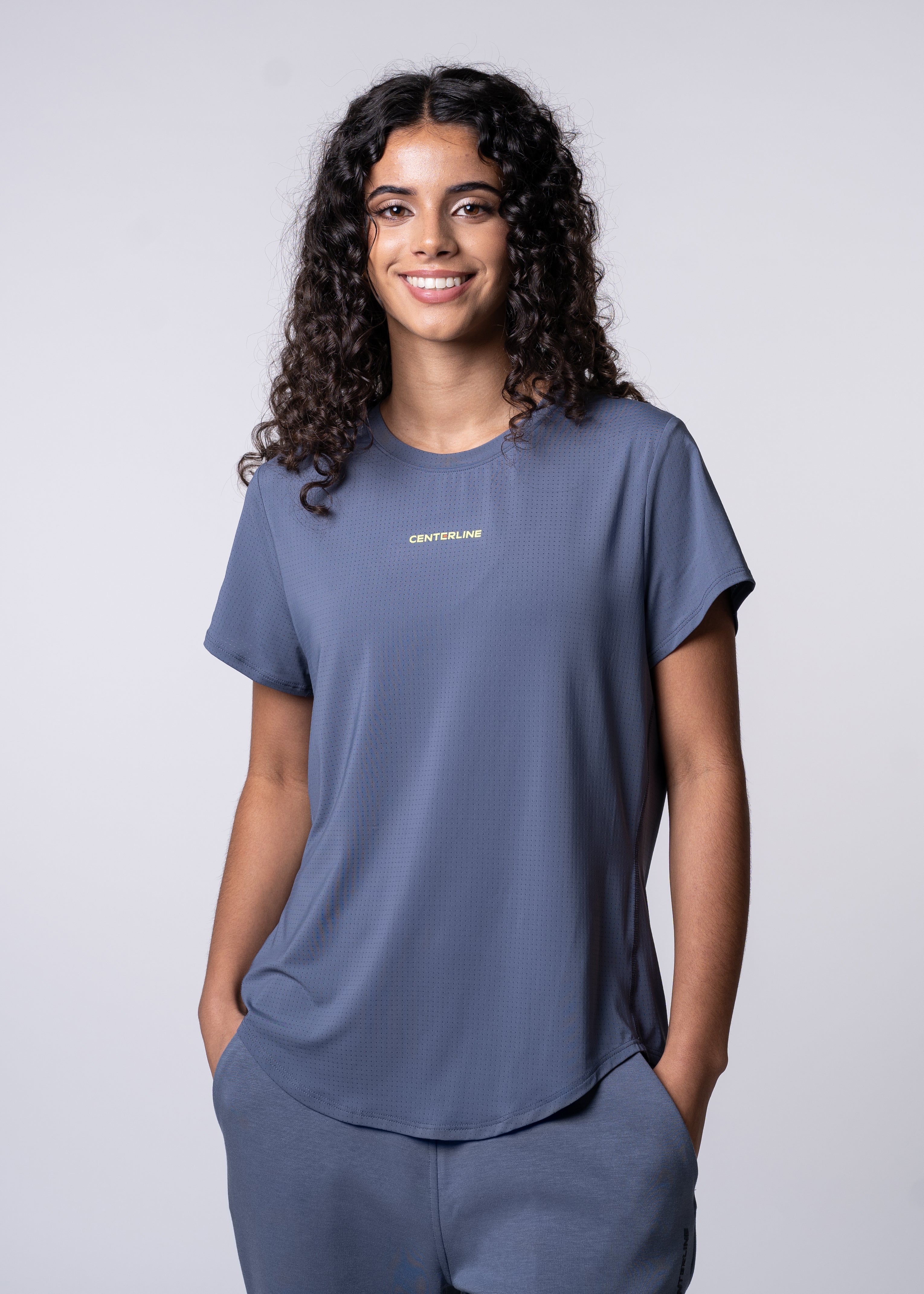 A woman in a blue athletic t-shirt and pants, smiling, with her hands in her pockets.