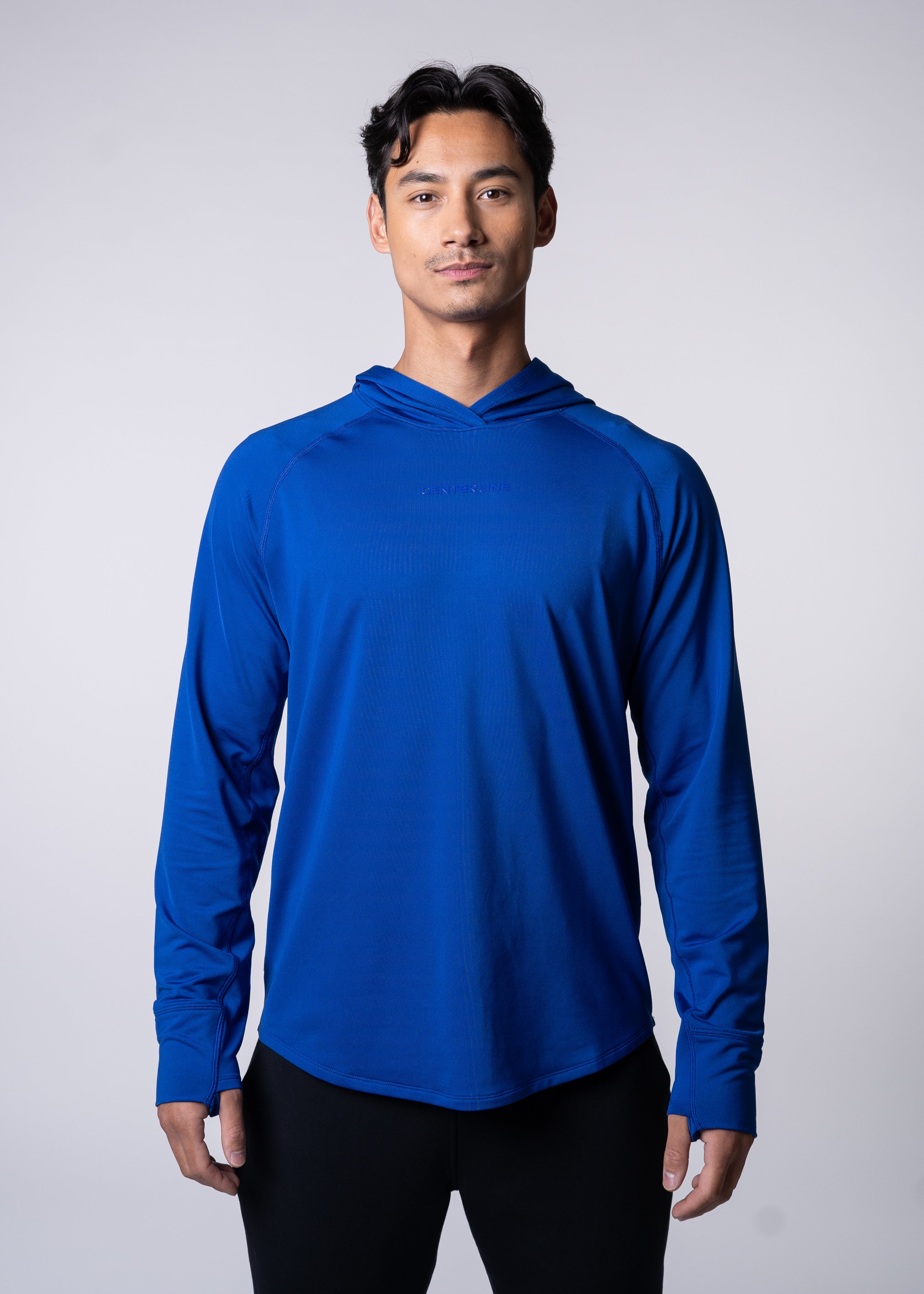 A man in a blue long-sleeve hooded shirt and black pants standing against a white background.
