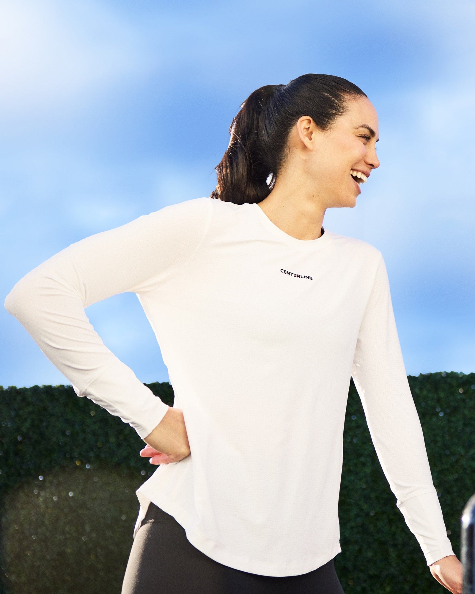 A woman in a white long-sleeve shirt and black leggings laughs outdoors.