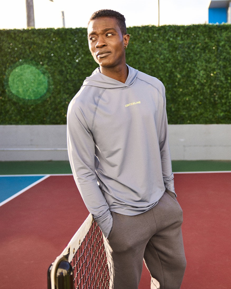Man wearing a grey hooded sweatshirt and sweatpants, standing on a tennis court.