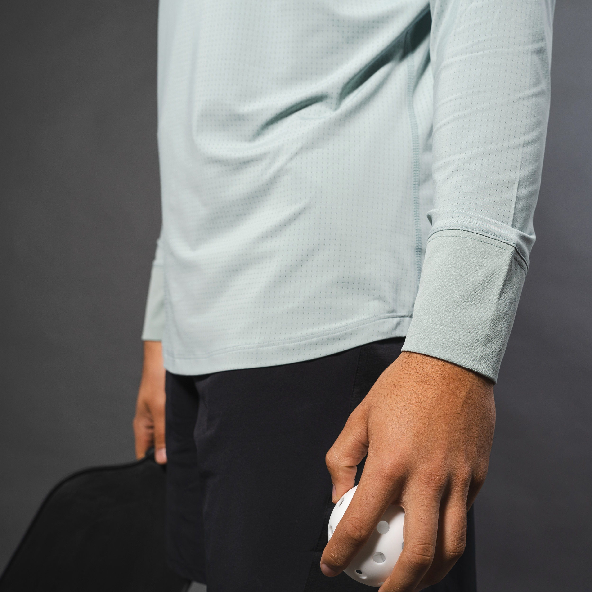 Person holding a white pickleball and a paddle. They are wearing a light blue long-sleeved shirt and black shorts. Gray background.