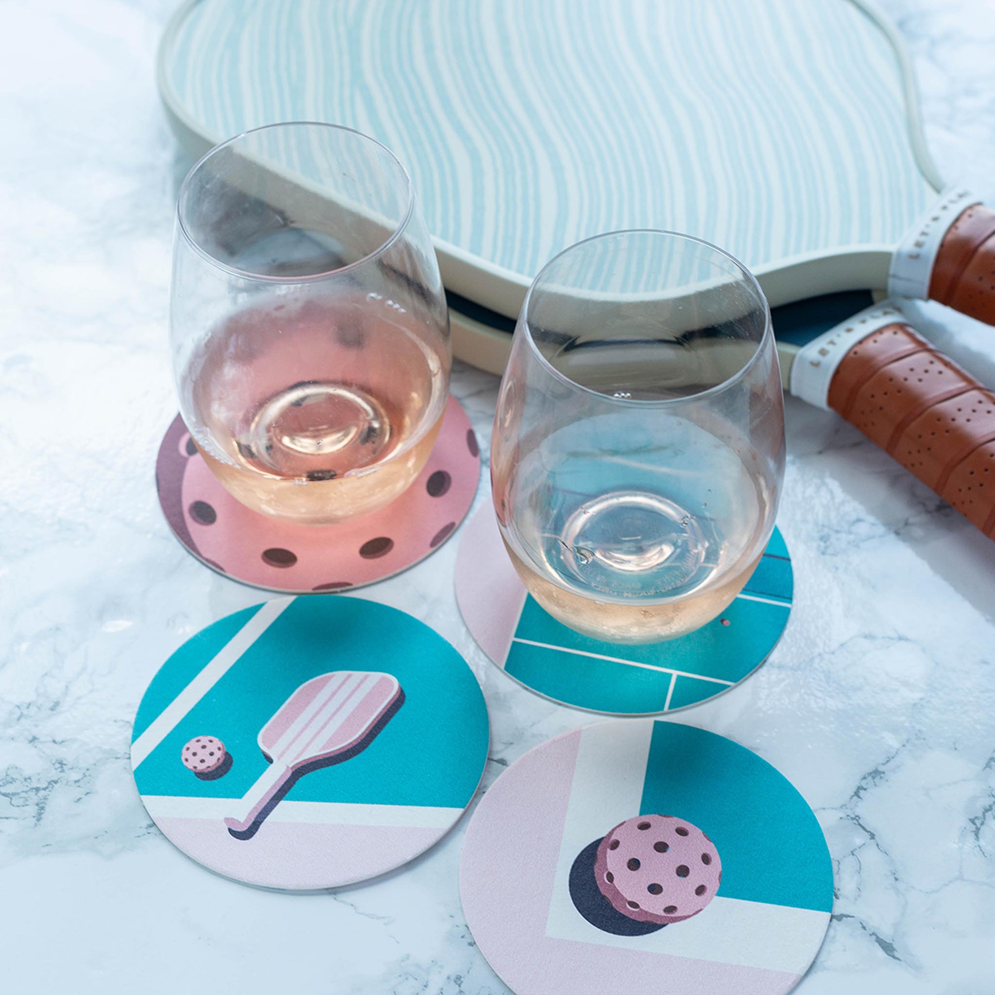 Two glasses of rosé wine on coasters with pickleball-themed designs, next to a pickleball paddle on a white marble surface.