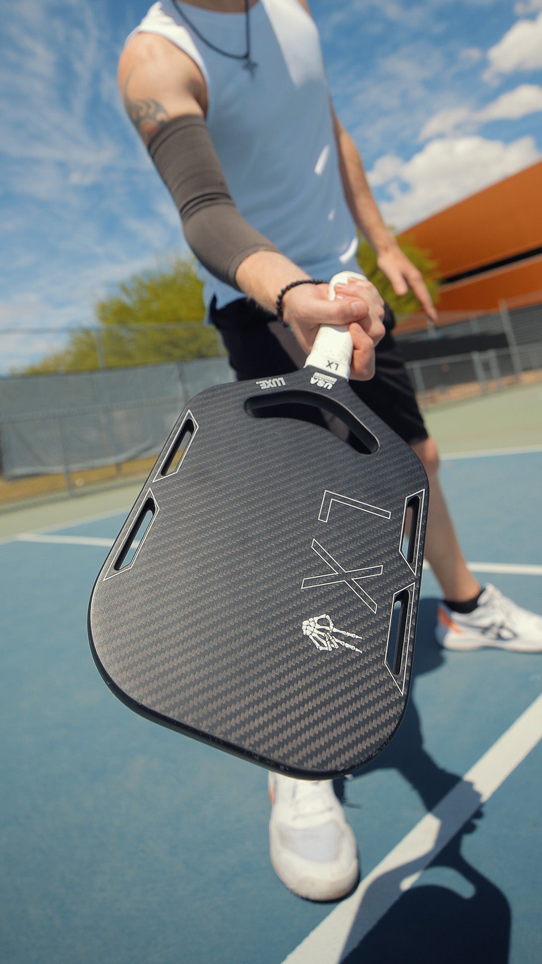 Person holding black and gray pickleball paddle on blue court, wearing white tank top and black shorts.
