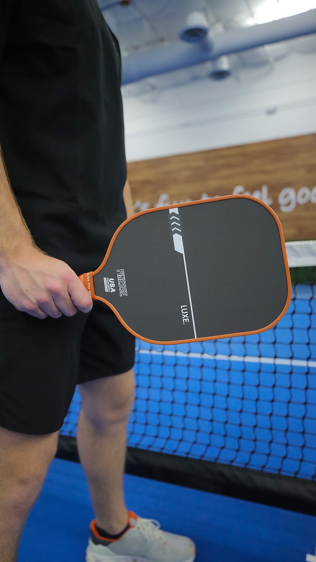 Man holding a Finesse Luxe pickleball paddle, black shorts, white sneakers, indoor pickleball court.