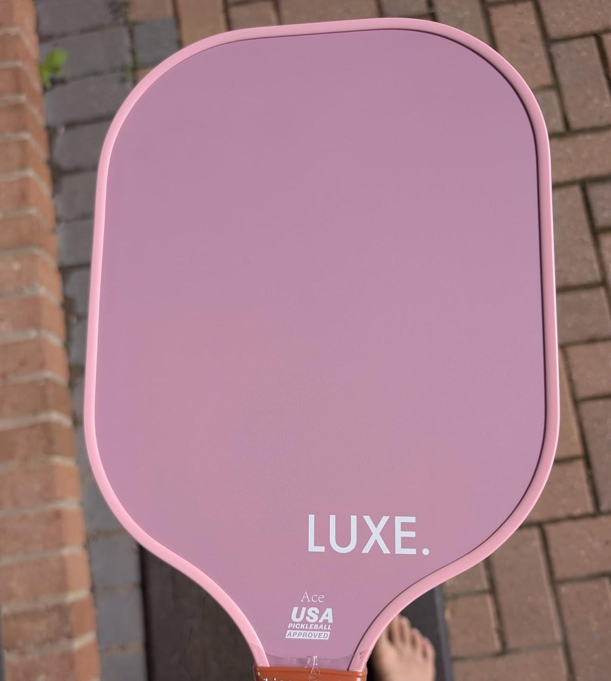 Mauve pickleball paddle with white "LUXE." text and USA Pickleball approval mark, shown from front view on brick patio.