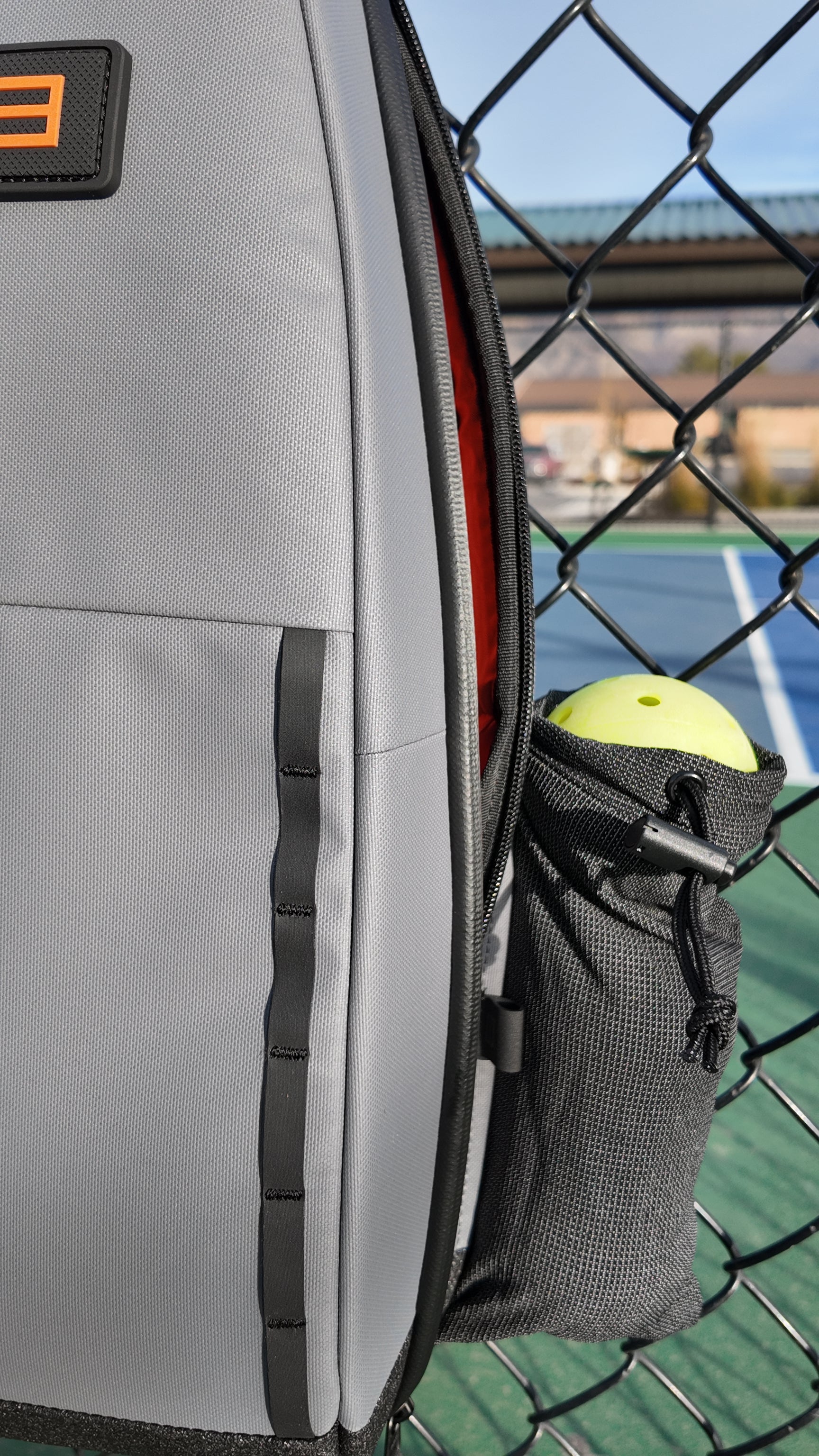 Gray pickleball sling by FORWRD with an orange E logo and a side pocket holding a yellow pickleball, next to a court fence.
