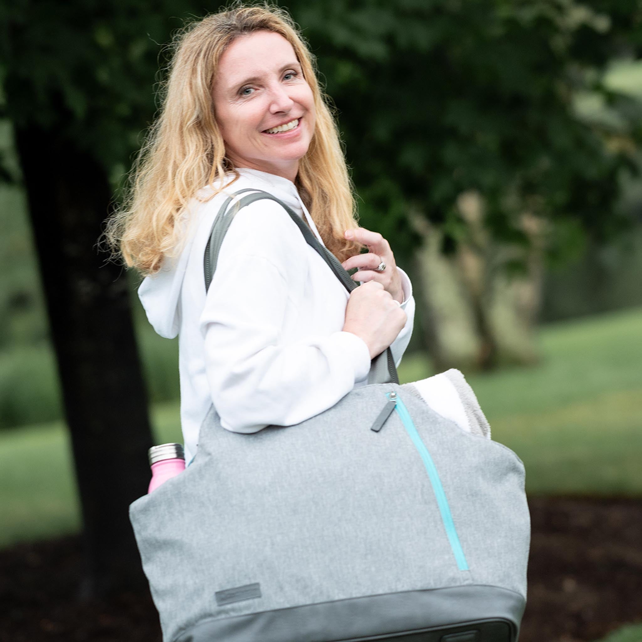 A blonde woman in a white hoodie carries a gray tote bag with a pink water bottle.