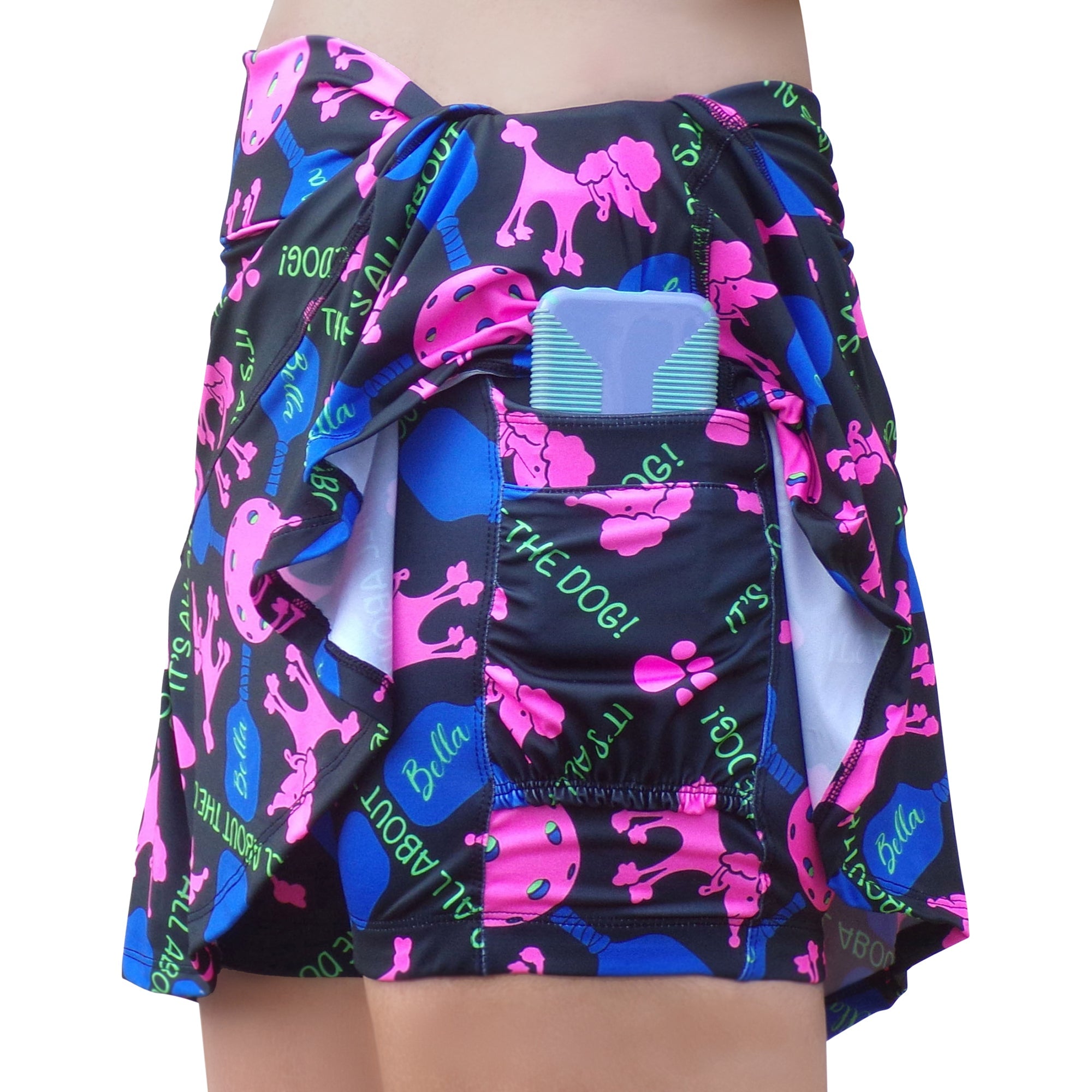 Black pleated skort with a pink, blue, and green pickleball and poodle print by Pickleball Bella, featuring "IT'S ALL ABOUT THE DOG!" text and a device in a pocket.