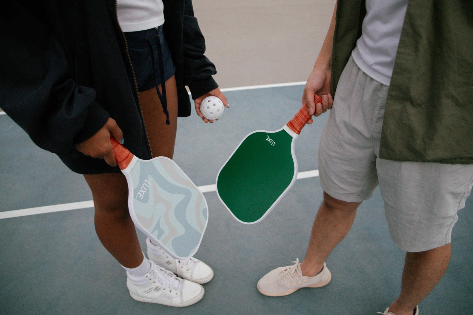 Two people holding pickleball paddles on a court, one person is holding a pickleball. The paddles are green and blue, and the people are wearing casual clothing and sneakers.