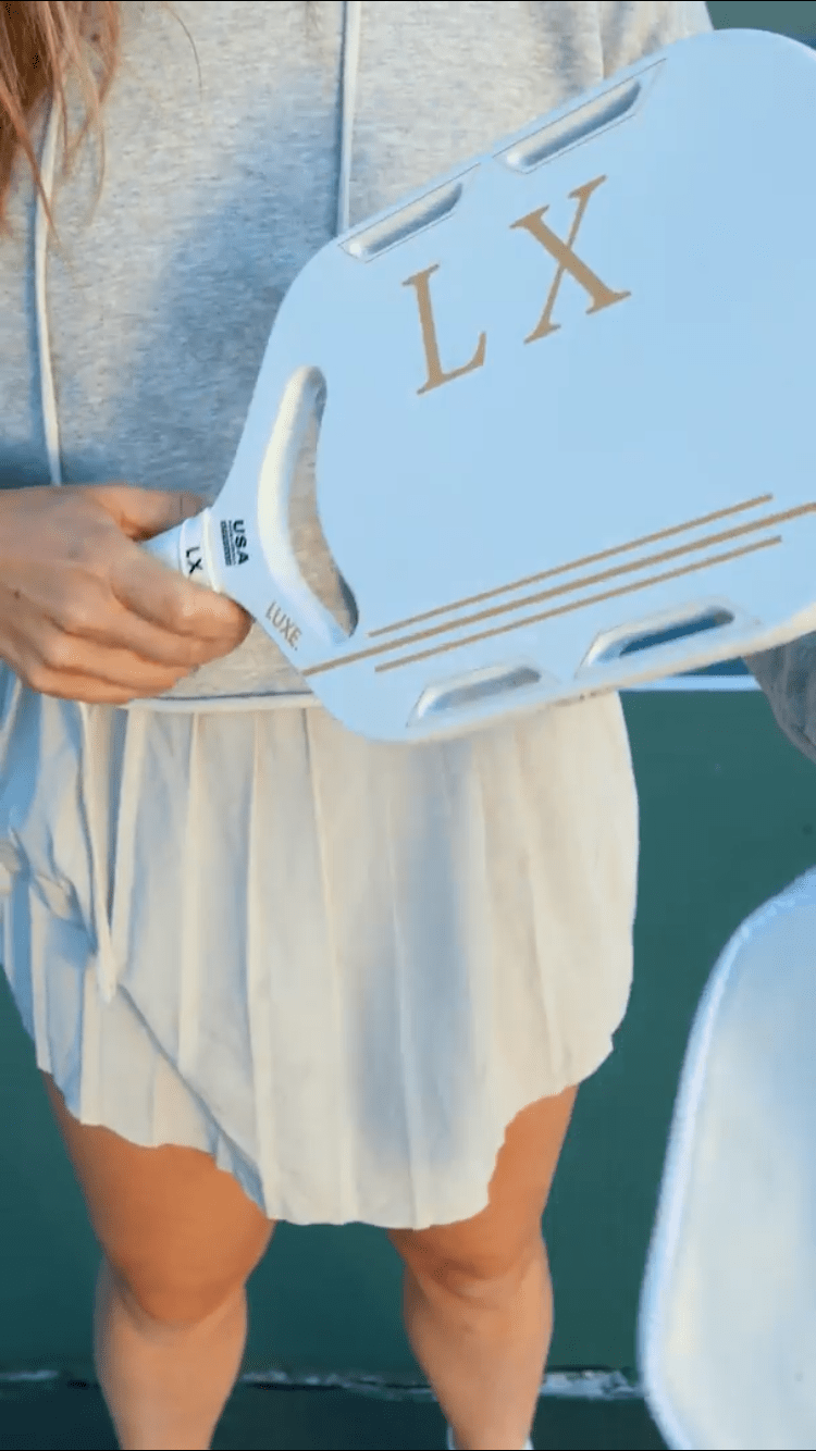 Woman holding a light blue paddle with gold "LX" lettering, wearing a gray hoodie and pleated skirt.