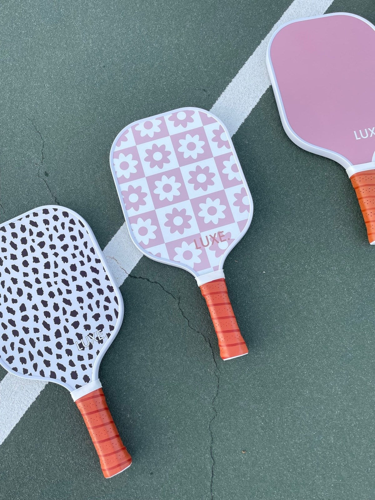 Three pickleball paddles with orange grips on a green tennis court.