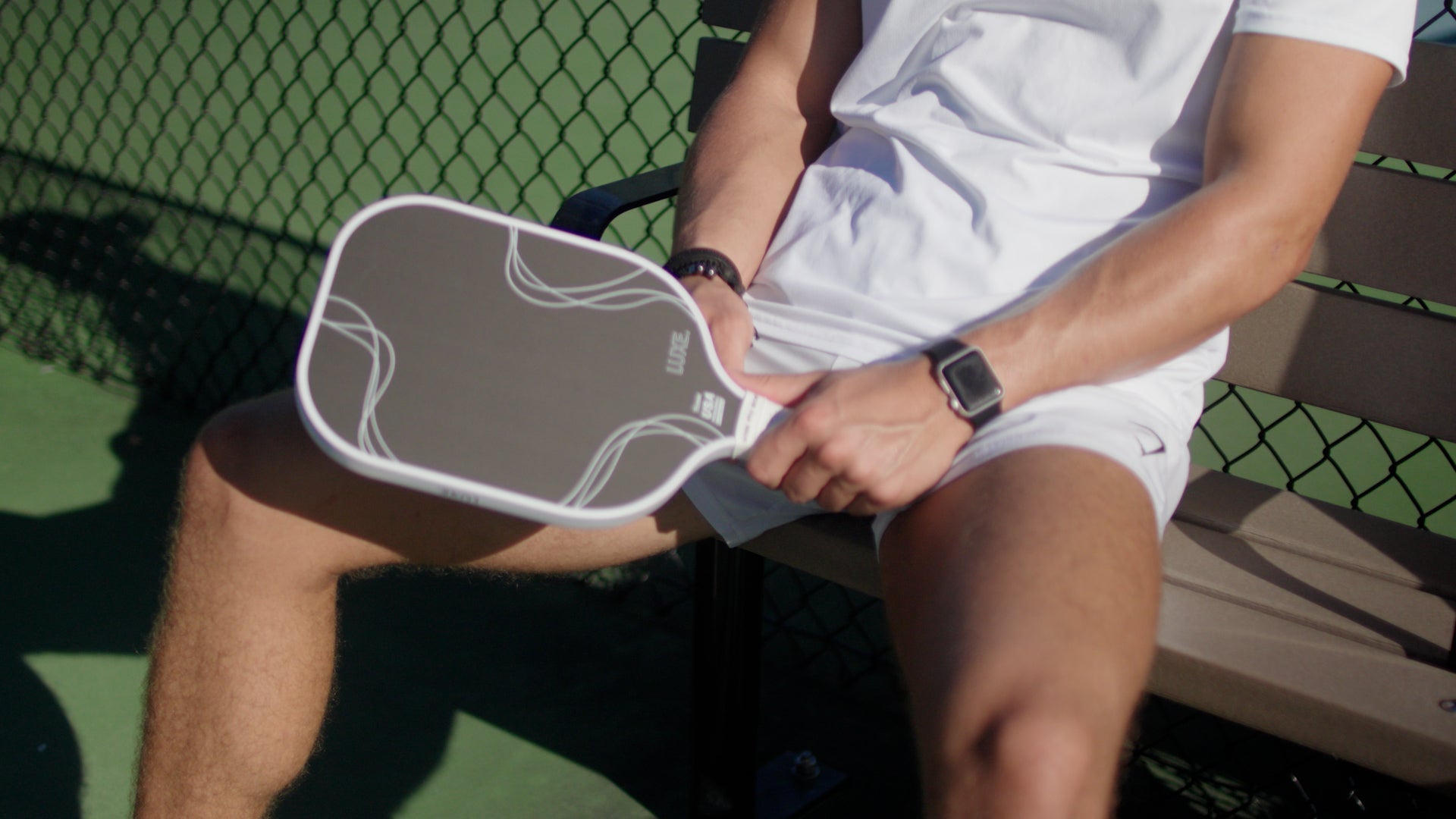 Man in white shorts and shirt, sitting on a bench, holding a pickleball paddle.