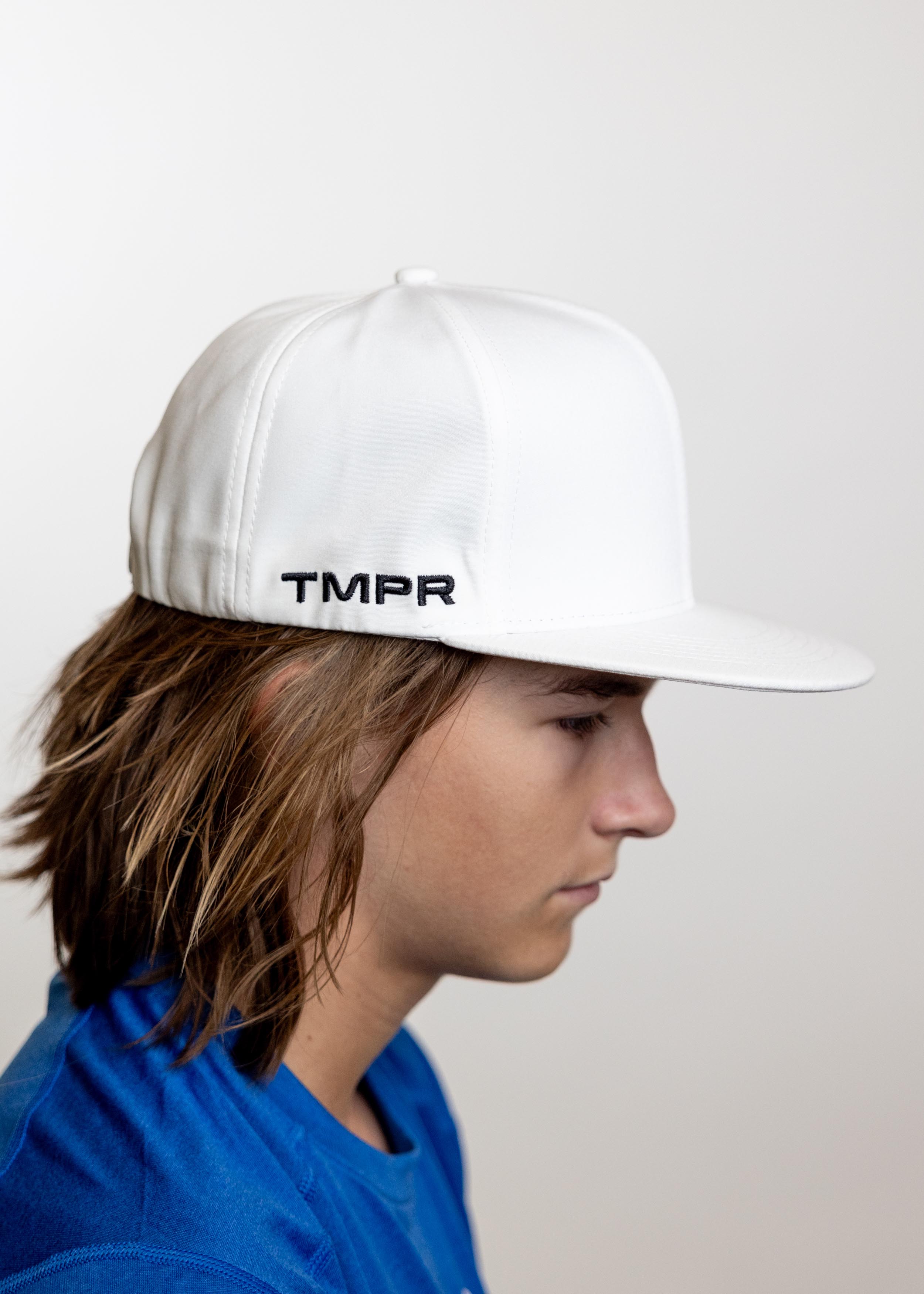 White flat bill cap by TMPR Sports with black 'TMPR' embroidery on the side, seen in a side view on a person wearing a blue shirt.