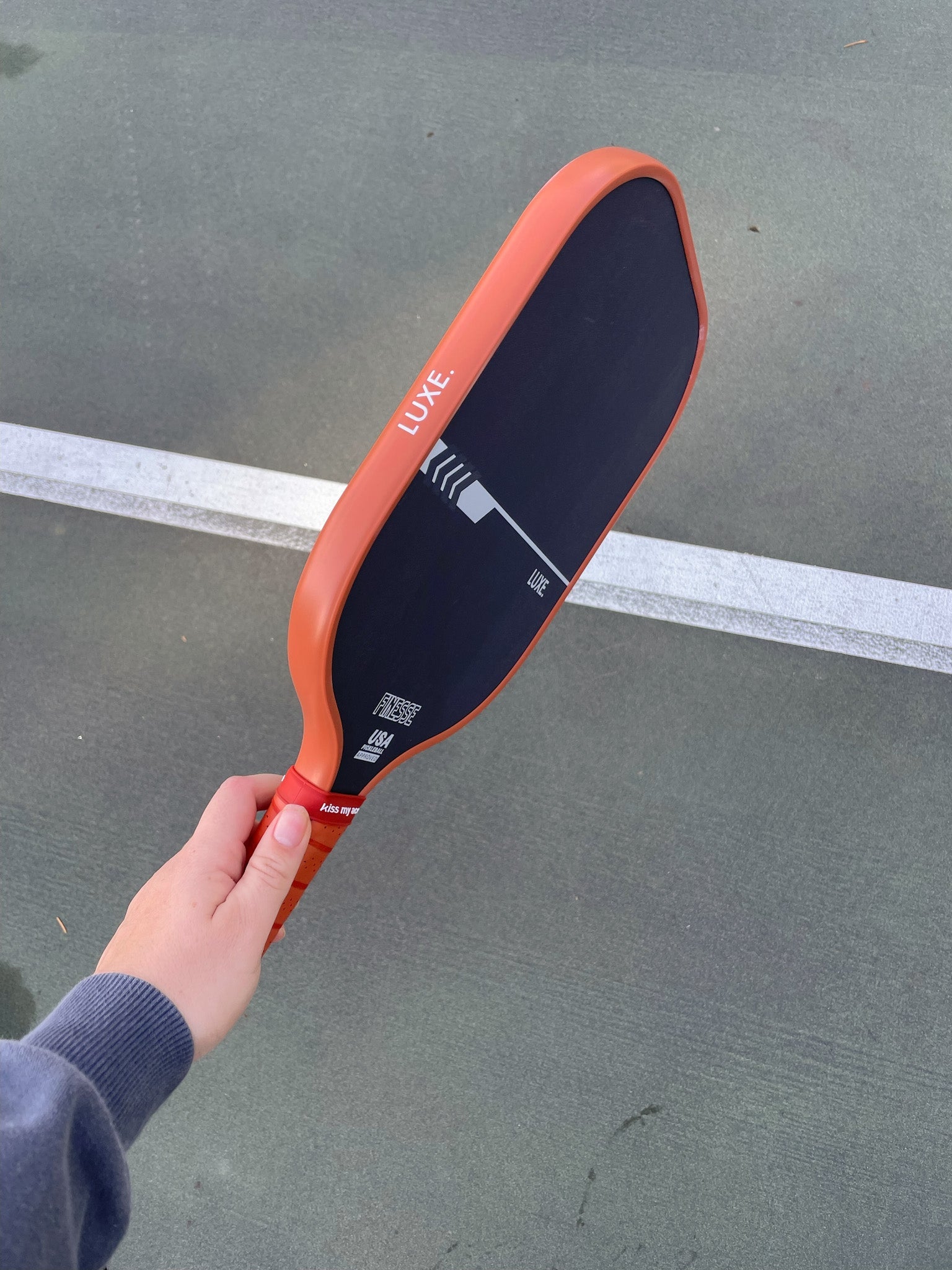 Hand holding a Luxe pickleball paddle on a grey court with a white line.
