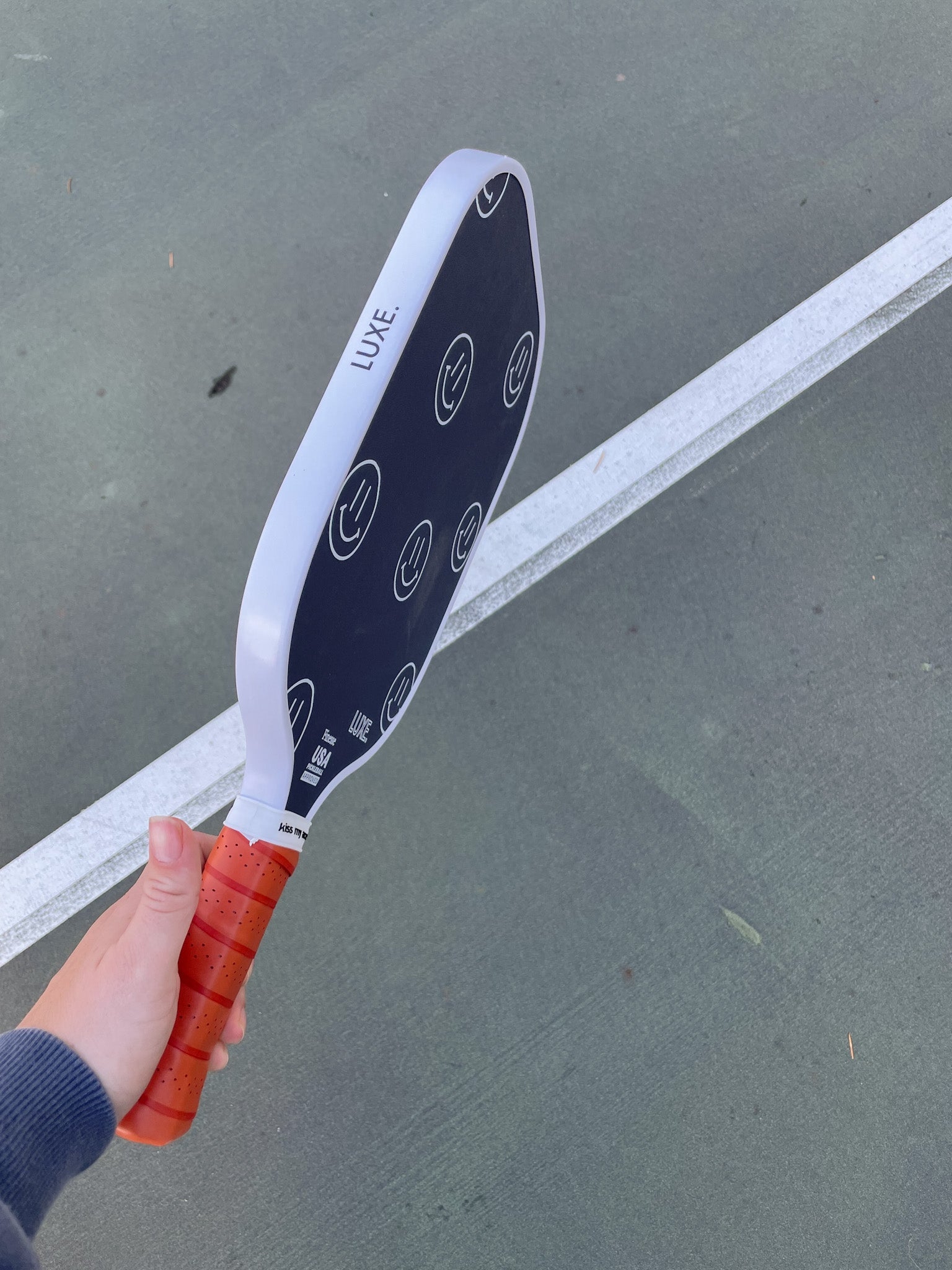 White and black Finesse pickleball paddle with an orange grip by LUXE Pickleball, featuring white smiley faces, held over a green court.