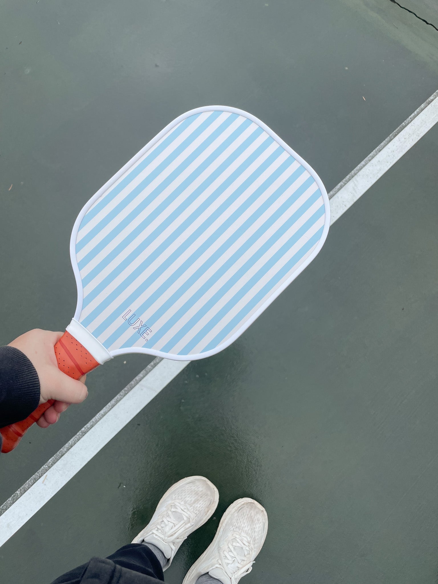 Person holding a striped pickleball paddle while standing on a tennis court wearing white sneakers.
