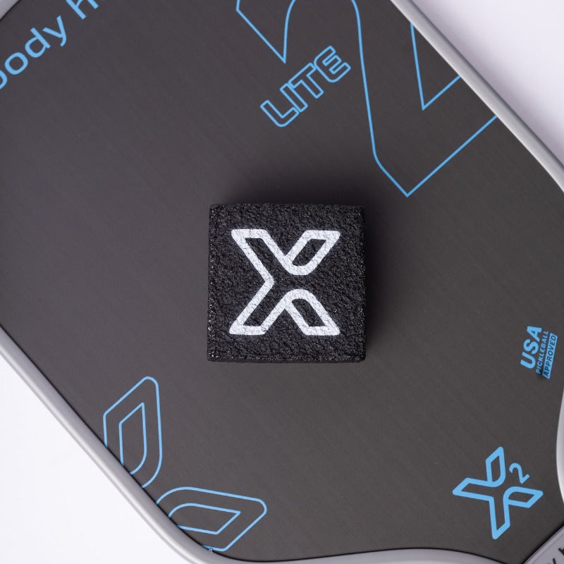 Close-up of a black pickleball paddle featuring a white "X" logo.