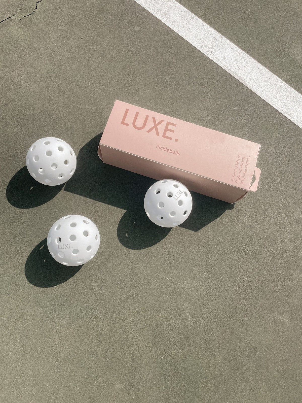 Three white pickleballs and a pink Luxe pickleballs box on a gray court with white lines.