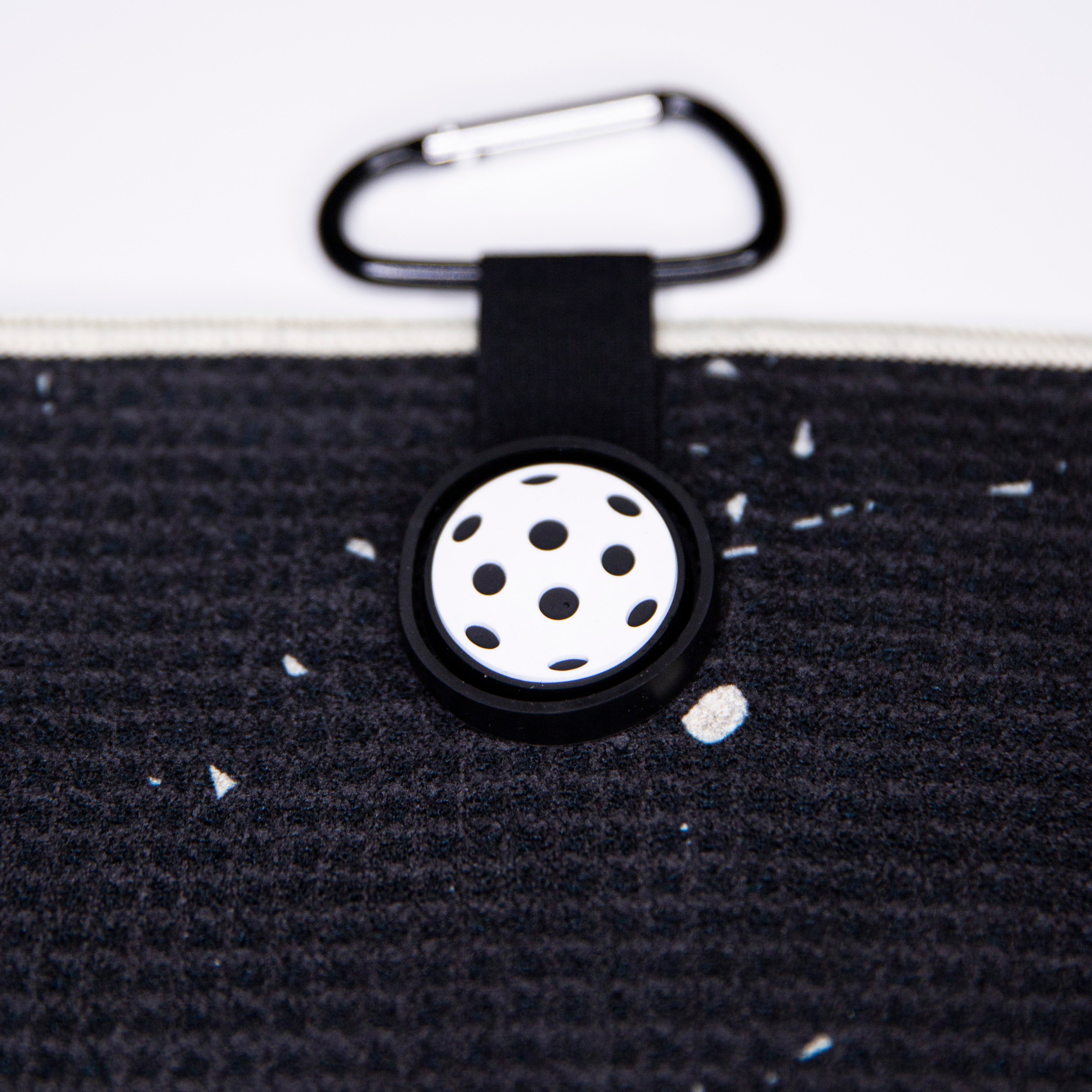 Black fabric with white flecks, black fabric loop, white and black pickleball design button, carabiner.
