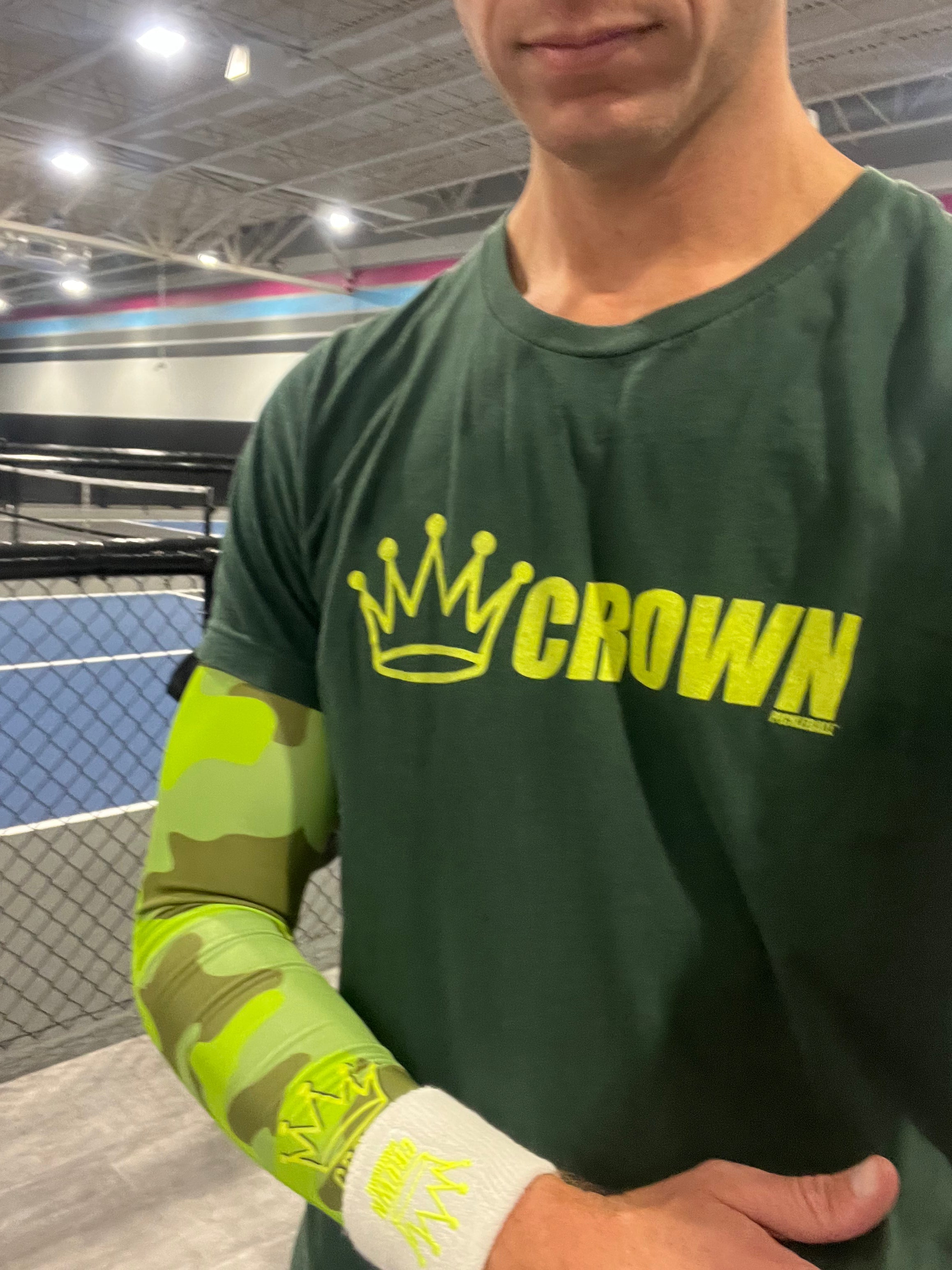 A man wearing a green T-shirt with a yellow "Crown" logo and a camouflage arm sleeve with a white wristband.