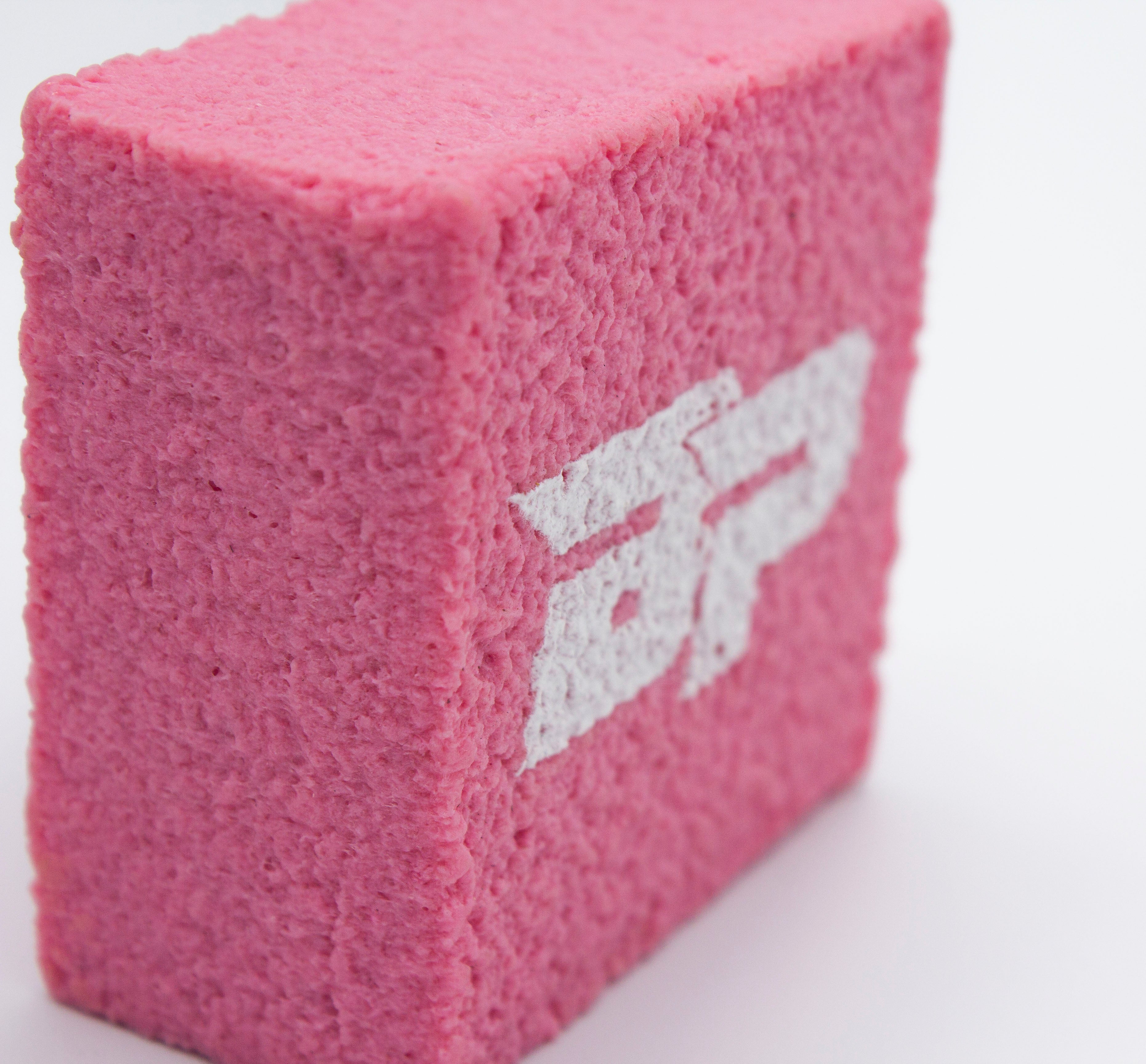 Pink, porous cube with white logo on white surface.
