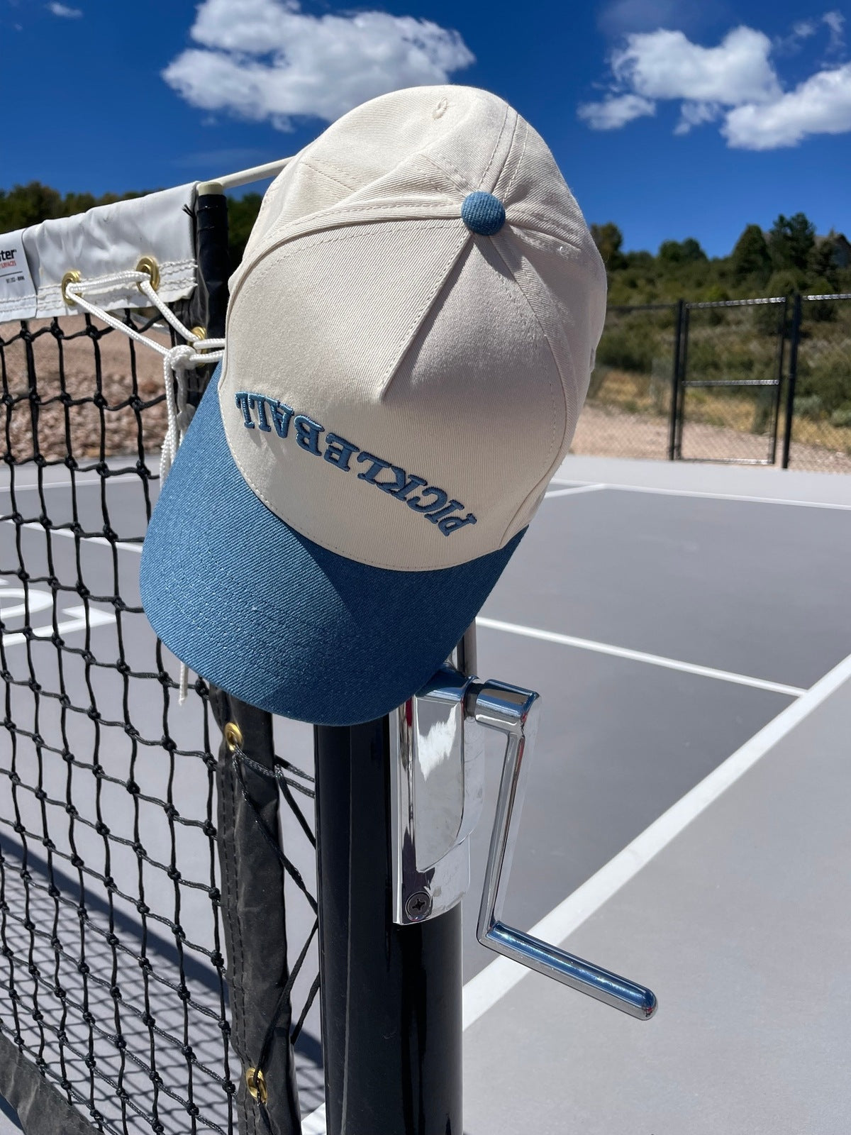 Beige and blue cap with "PICKLEBALL" embroidered on the side, hanging on a pickleball net.
