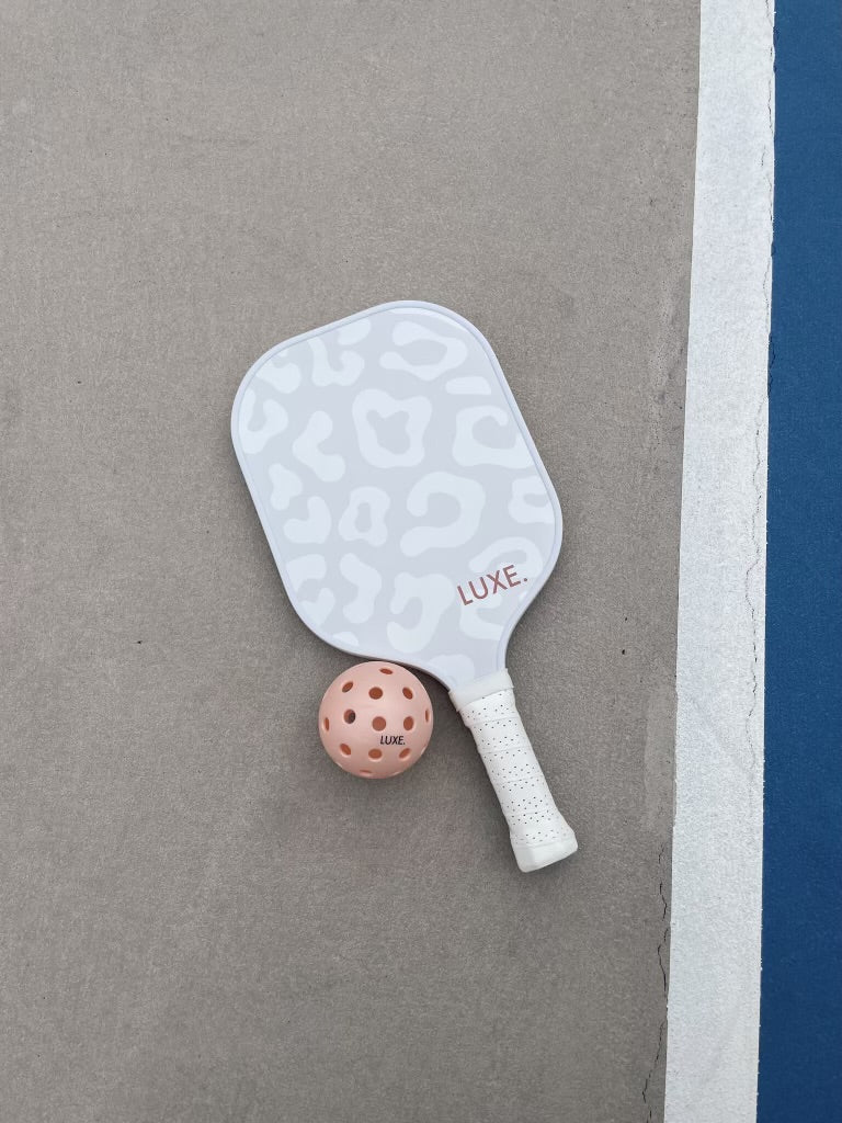 A white and light gray cheetah print pickleball paddle and peach pickleball with the word "LUXE" on both on a tennis court.