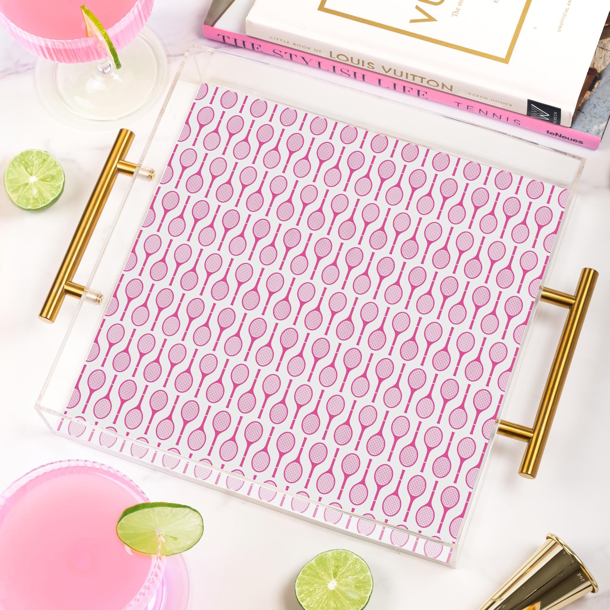 A clear serving tray with gold handles, decorated with a pink tennis racket pattern, resting on a white marble surface with drinks, lime wedges, and books.