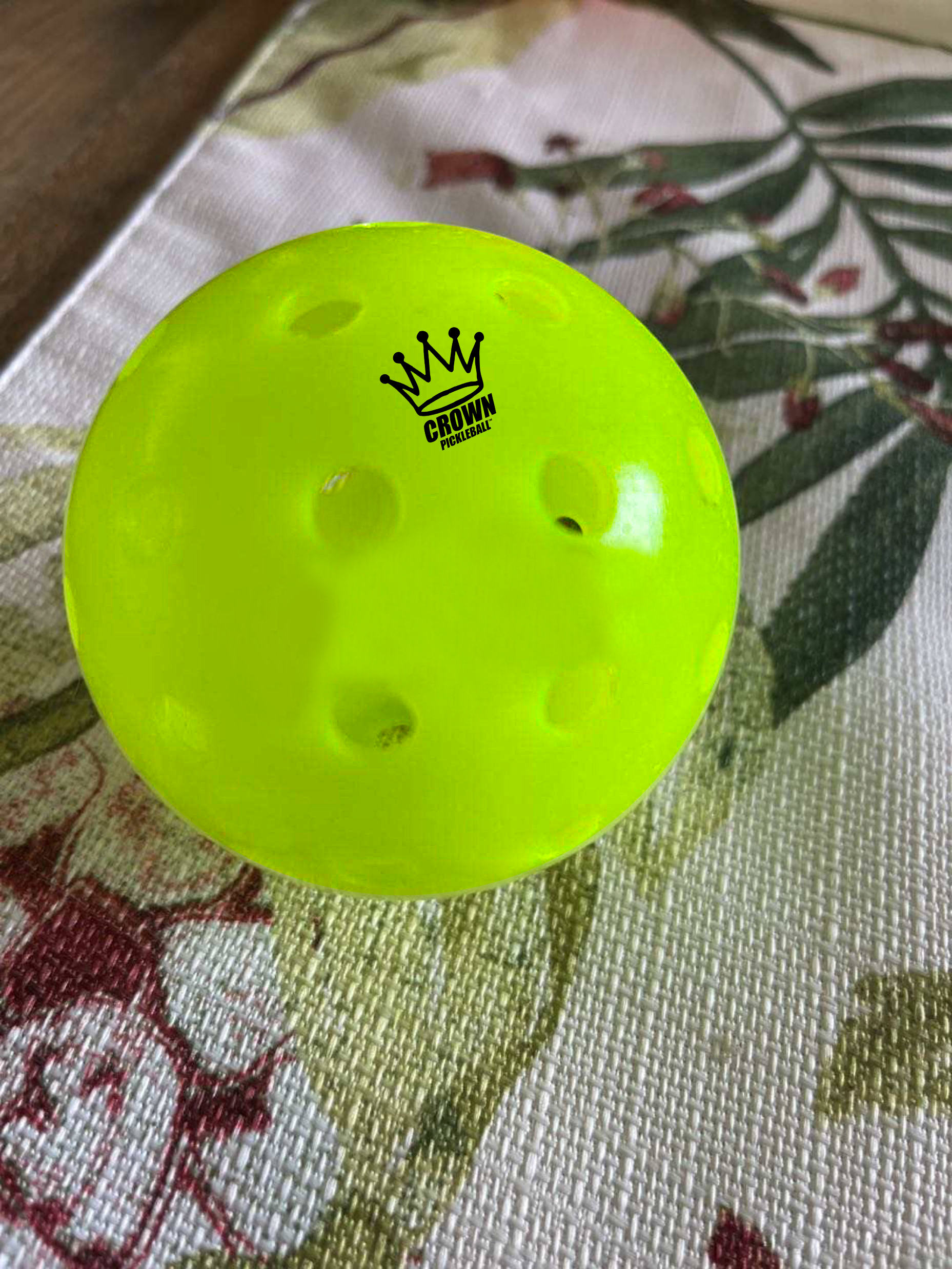 Close-up of a bright green pickleball with black logo on a textured, floral-patterned surface.