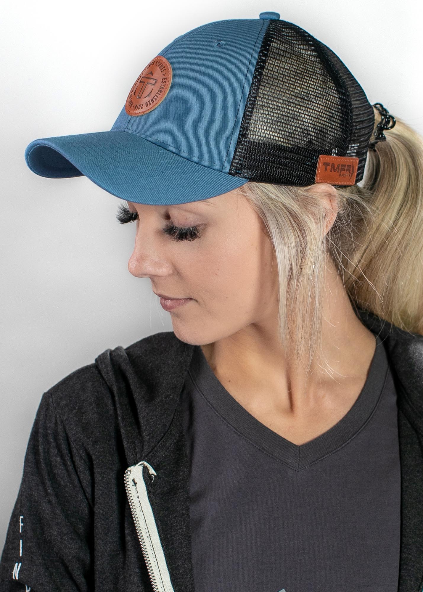 Blue trucker cap by TMPR Sports with a T logo leather patch on the front and TMPR tag on the mesh back, worn by a woman.