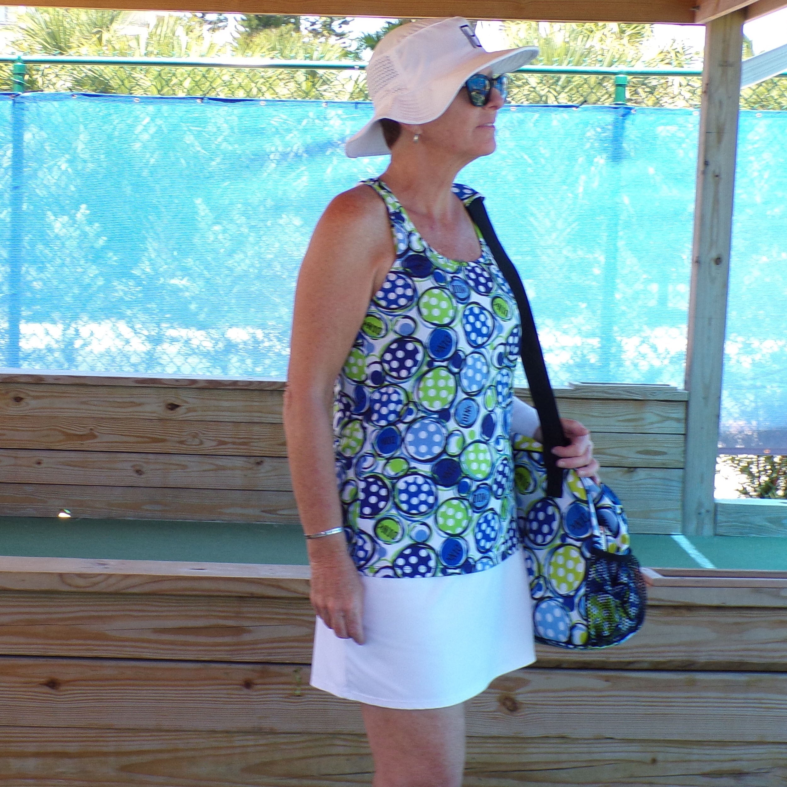 White, blue, and green polka dot Racerback Tank by Pickleball Bella, worn by a woman with a white skirt, visor, sunglasses, and a matching bag.