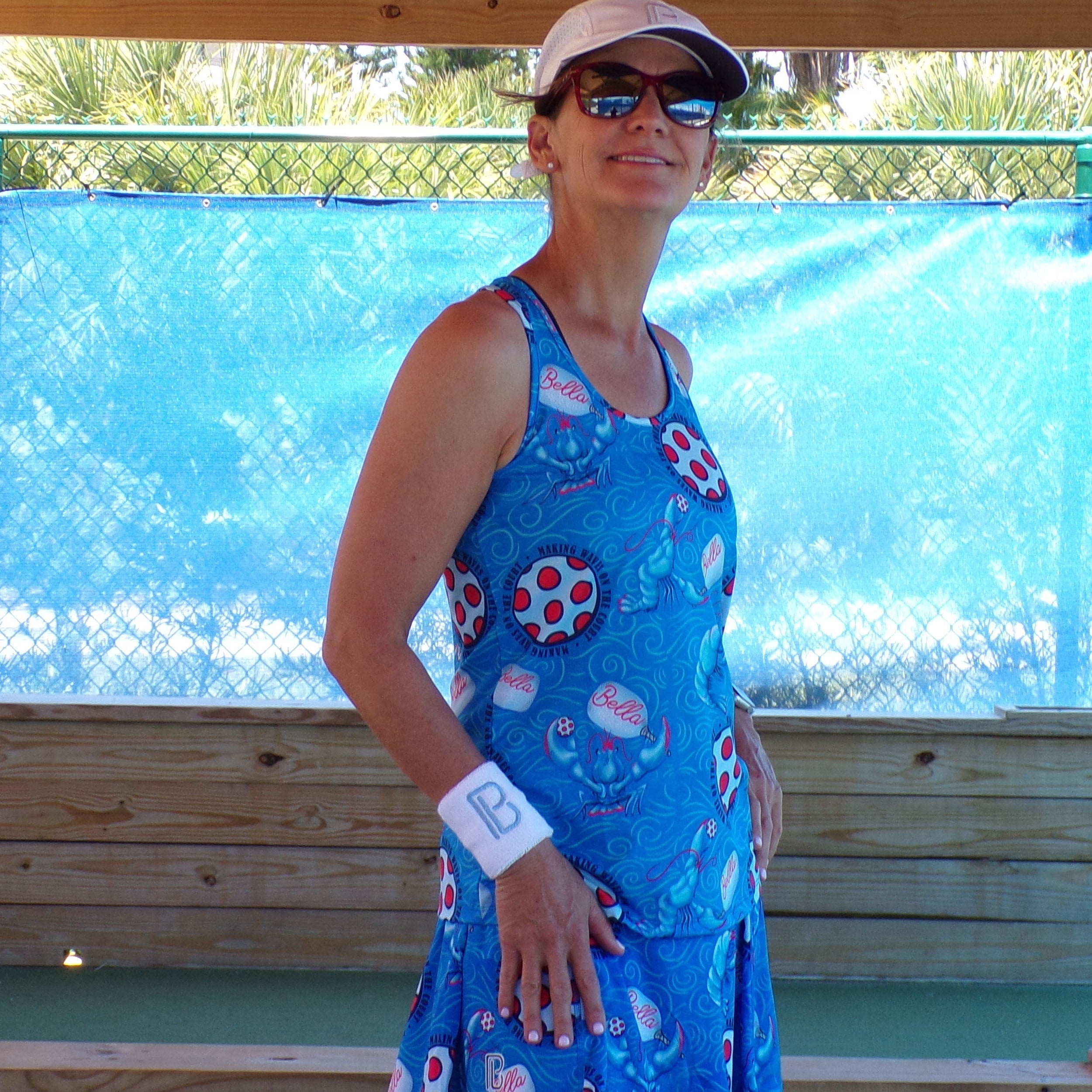 Blue Racerback Tank by Pickleball Bella with a Lobster Monster pattern, worn by a woman with a white visor, sunglasses, and a 'B' wristband.