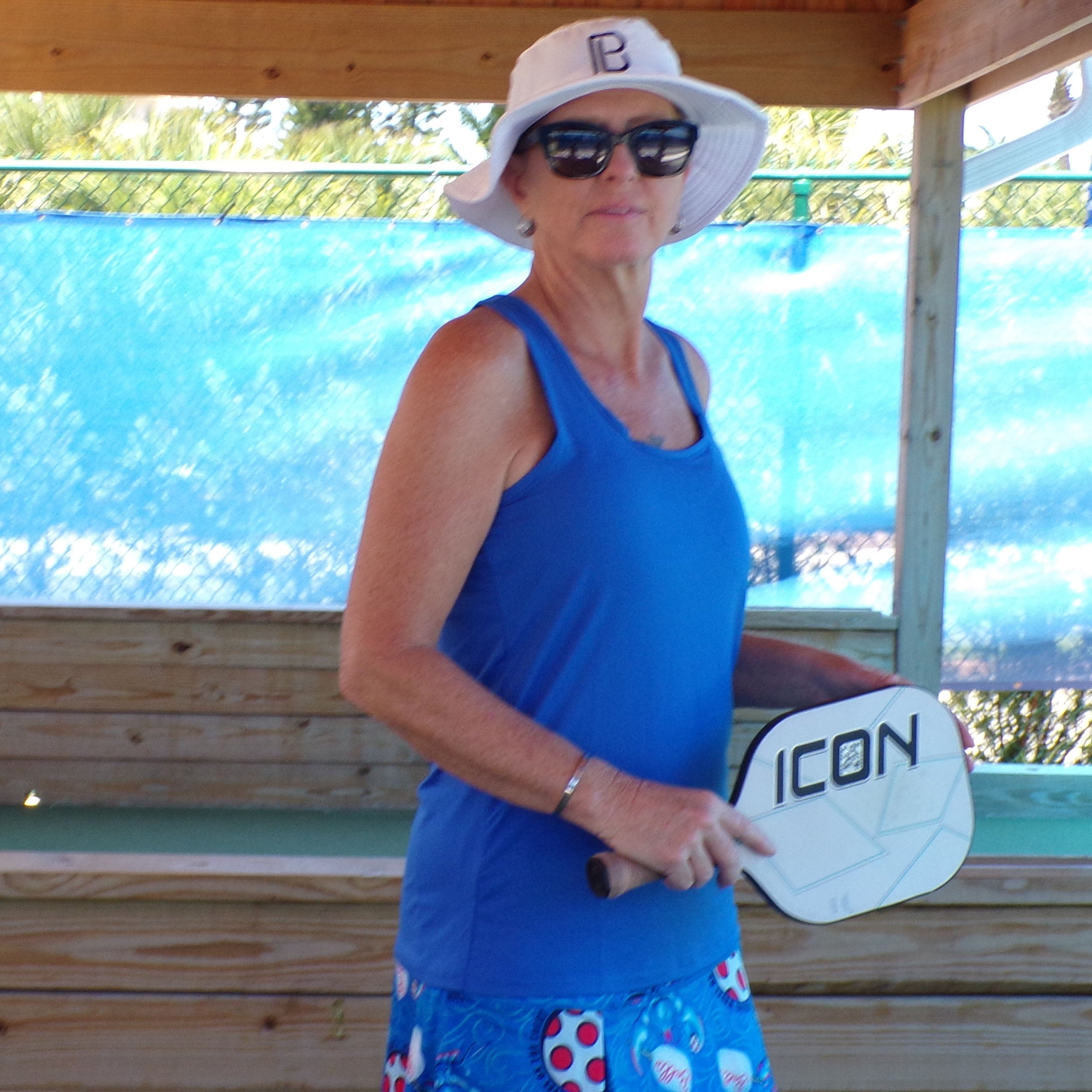 Royal blue racerback tank by Pickleball Bella, worn by a woman in a white hat, sunglasses, and a blue patterned skirt, holding a pickleball paddle.