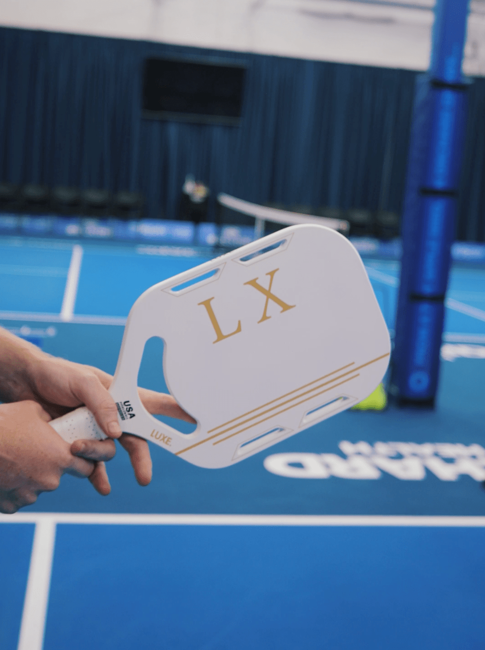 Person holding a white paddle with gold lettering in a blue indoor pickleball court.