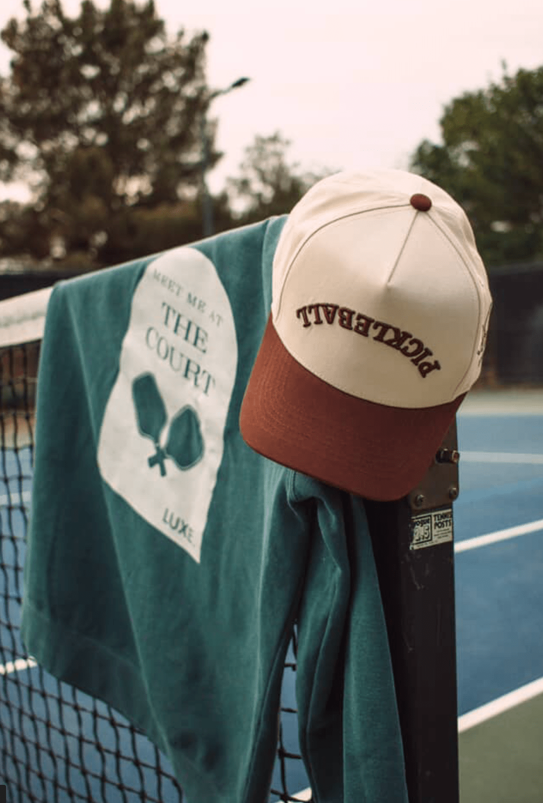 Product image of a green sweatshirt with a white logo hanging on a tennis net, next to a beige and brown baseball cap.