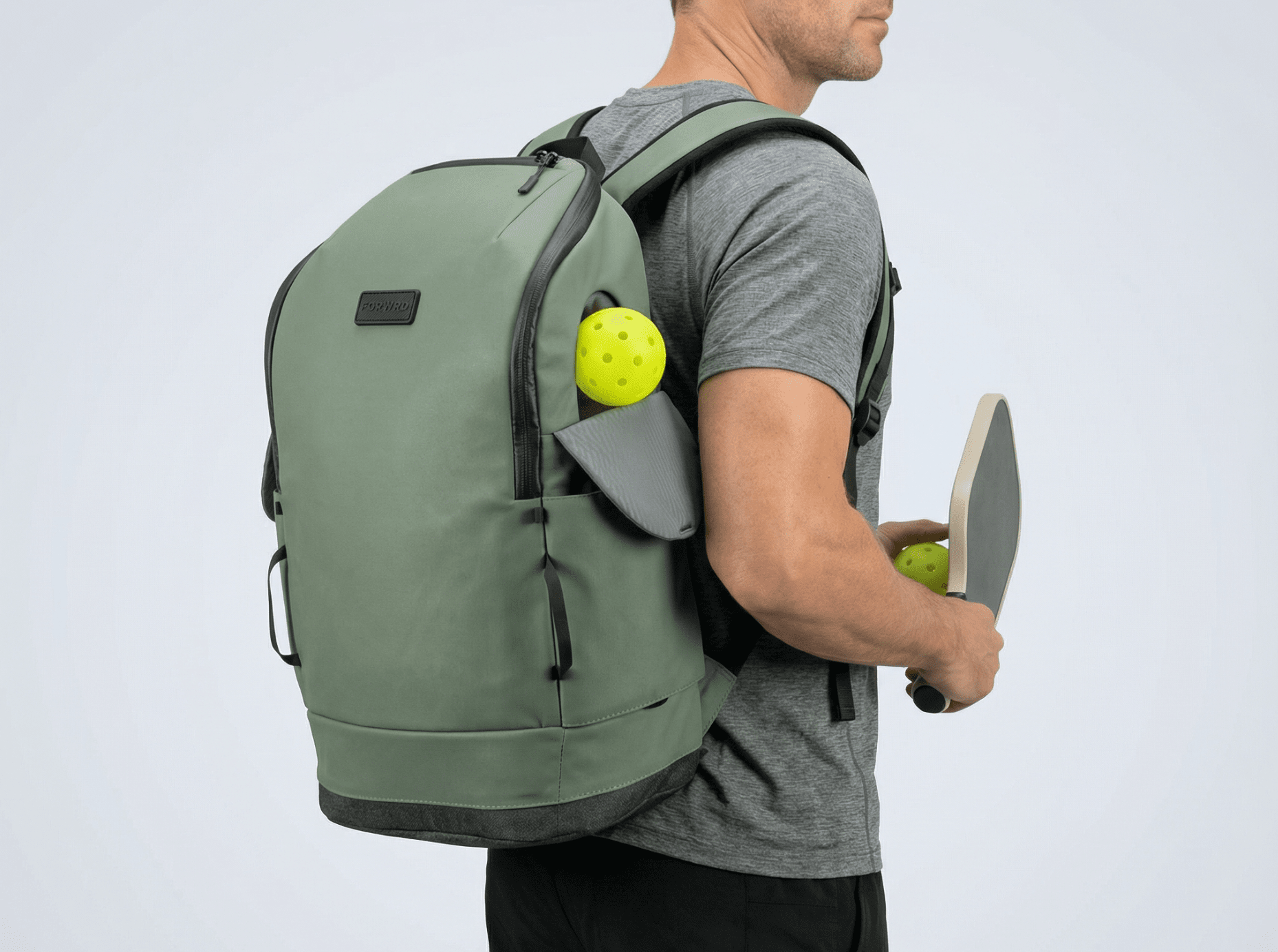 Sage green pickleball backpack by FORWRD with black accents and a yellow pickleball in the side pocket, worn by a person holding a paddle.