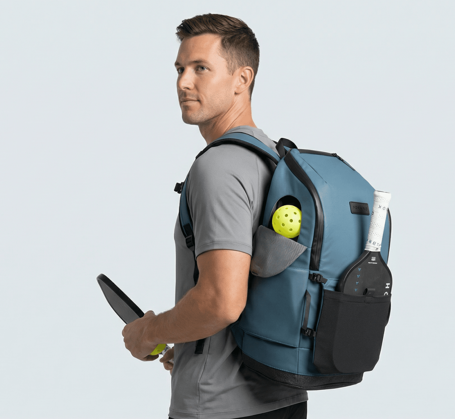 Blue pickleball backpack by FORWRD, worn by a man, with a pickleball and paddles in side pockets.