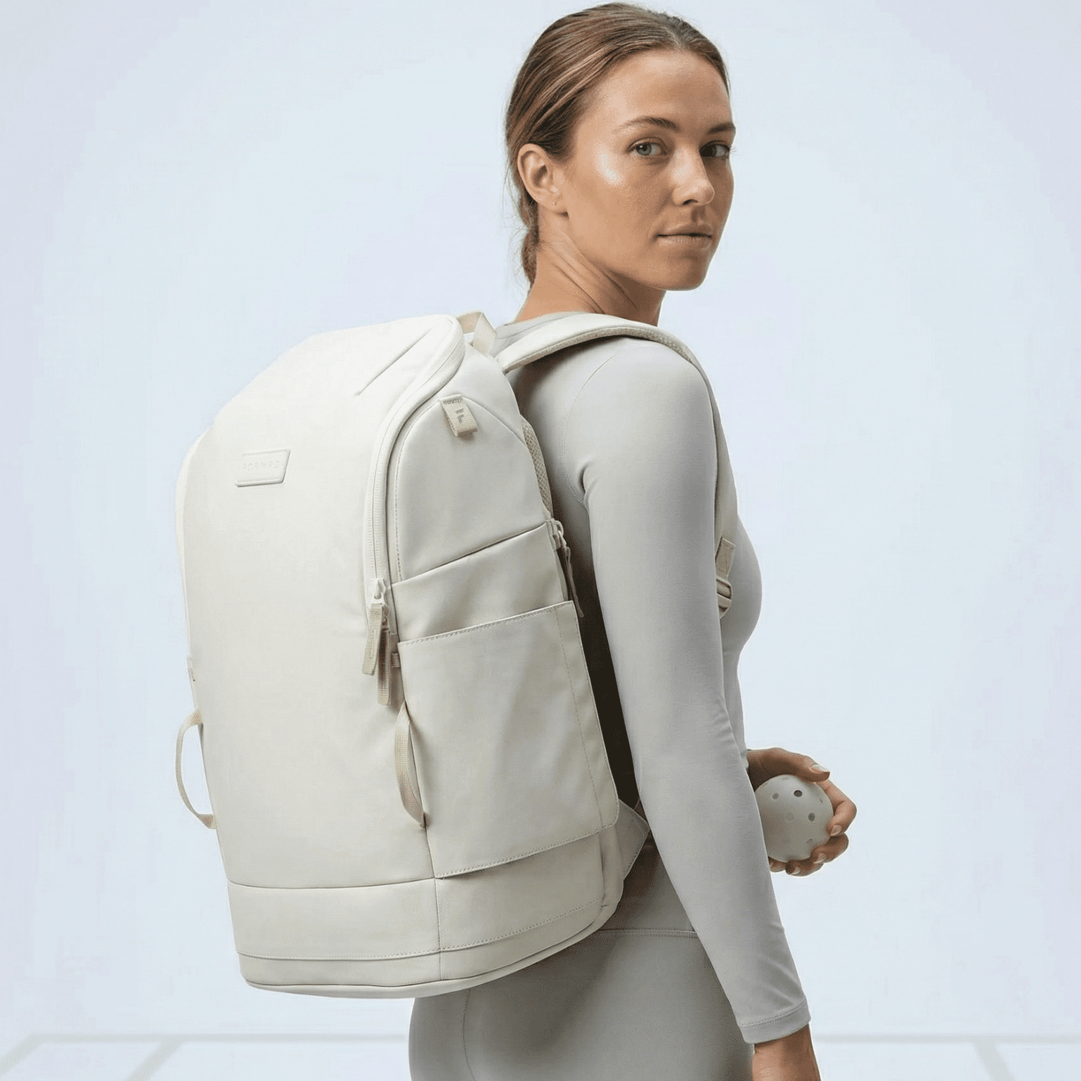 Light beige pickleball backpack by FORWRD, featuring zippered pockets, worn by a woman holding a white pickleball.