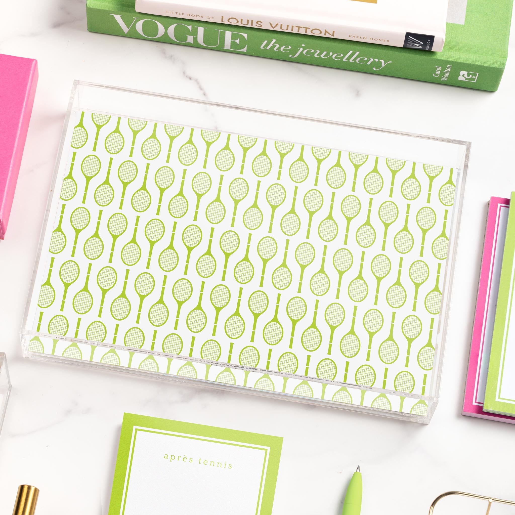 A clear tray with a tennis racket print is centered among books, a notepad, and a pen.