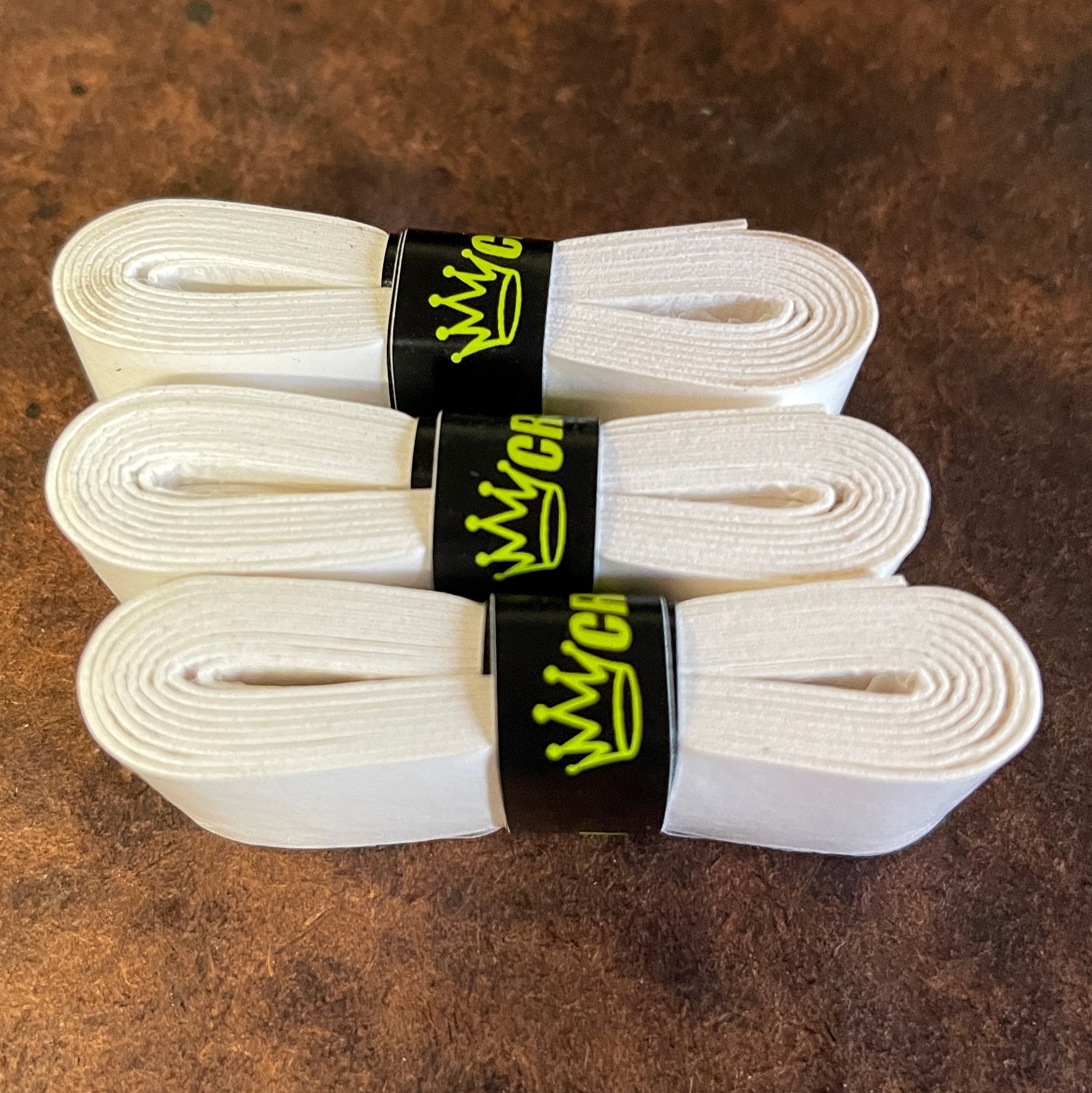 Three rolls of white grip tape with black labels and neon yellow crown logo, stacked on a textured brown surface.