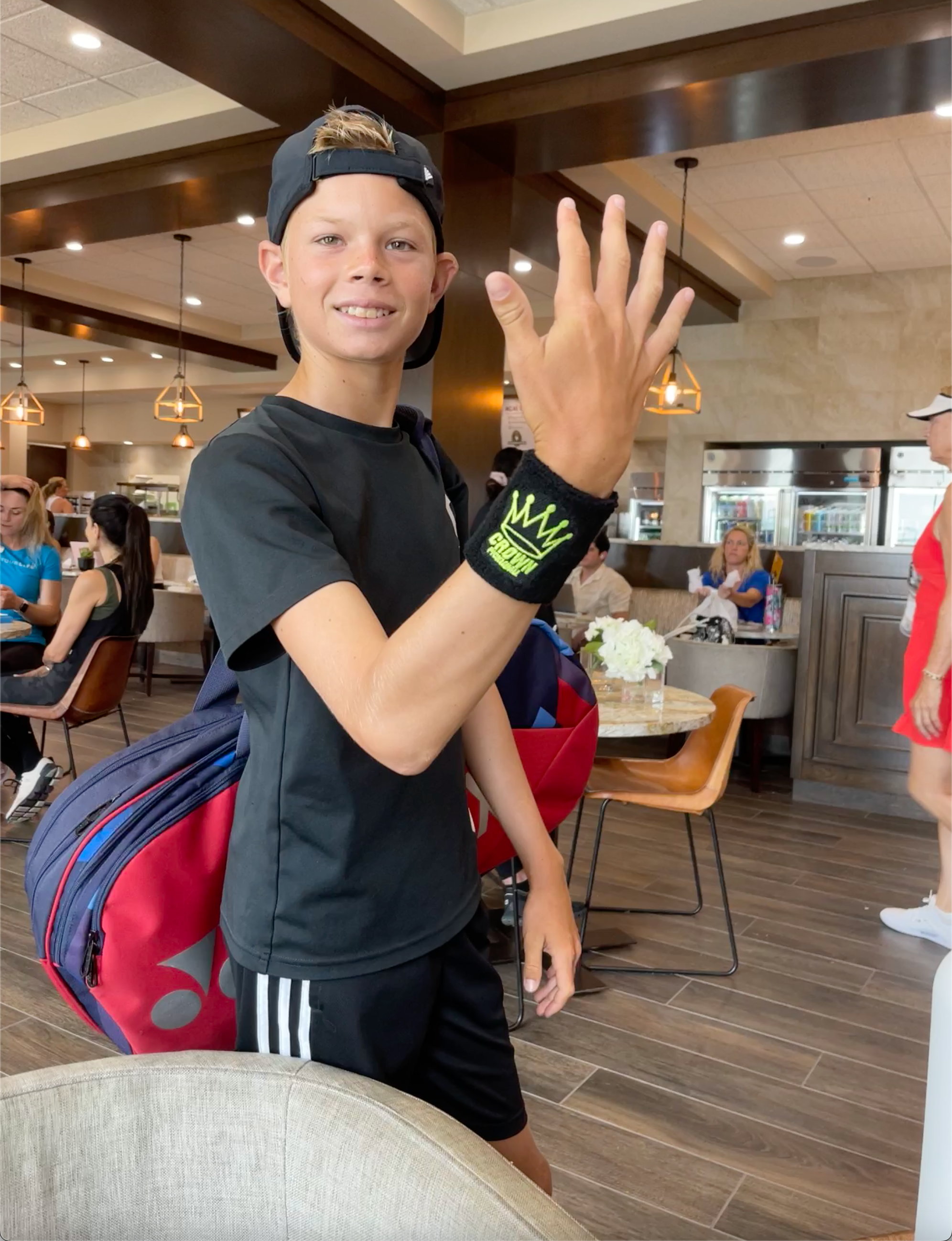 A young man in black athletic wear raises his hand while carrying a tennis bag.