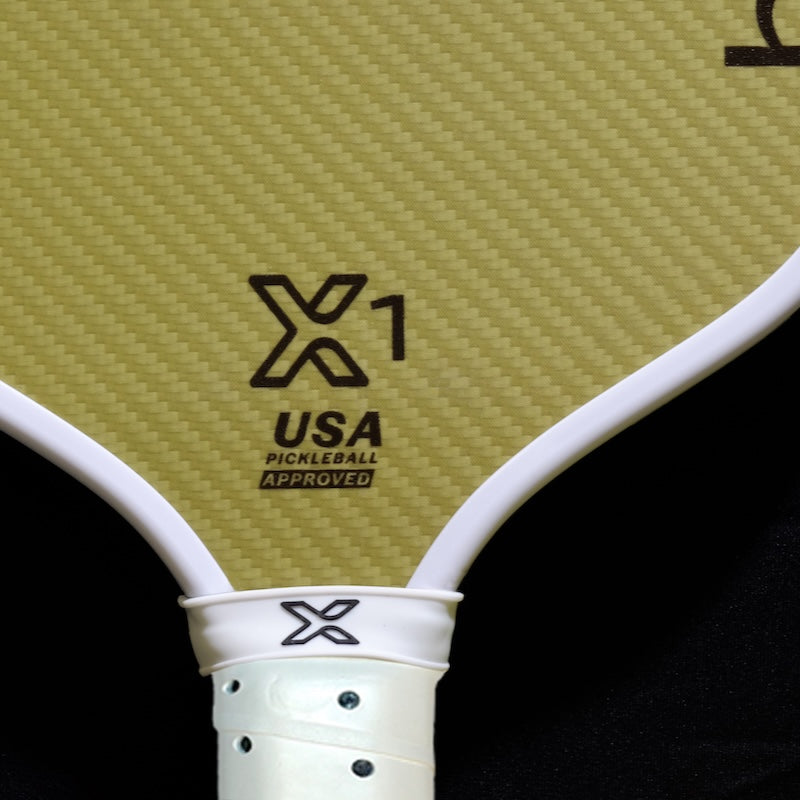 Close-up of a pickleball paddle with a woven texture, featuring the "X1" logo, "USA" text, and the phrase "PICKLEBALL APPROVED." The paddle has a white edge and a white grip with black accents.