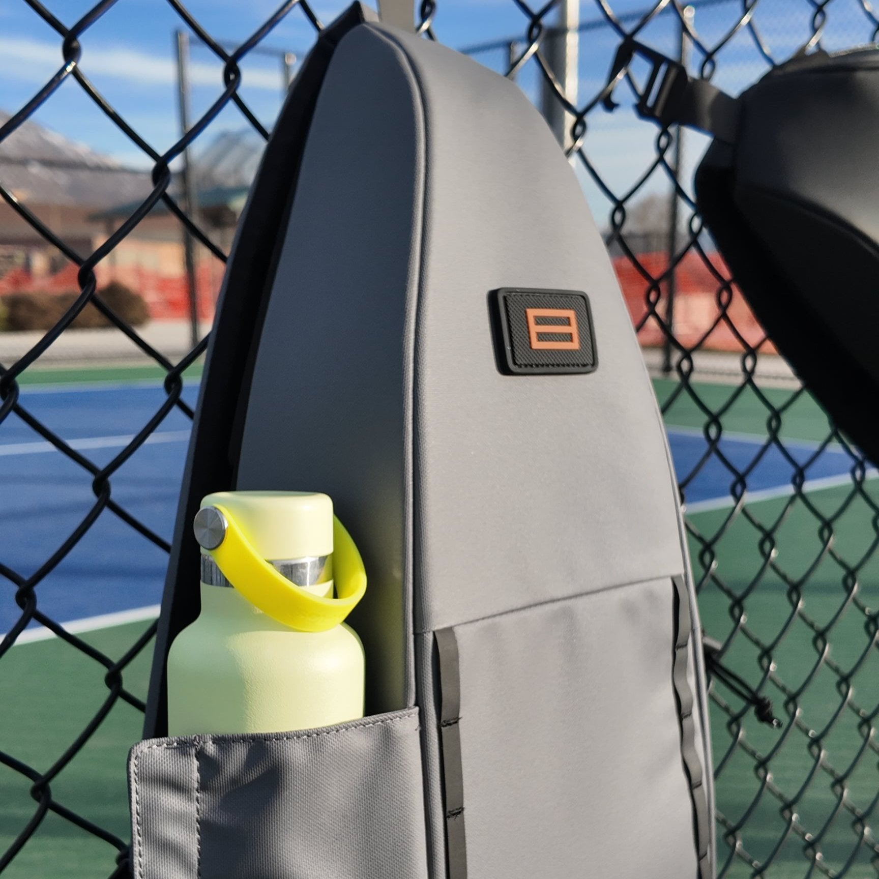 Gray Pickleball Sling by FORWRD, with an orange logo and a light green water bottle in a side pocket, hanging on a court fence.