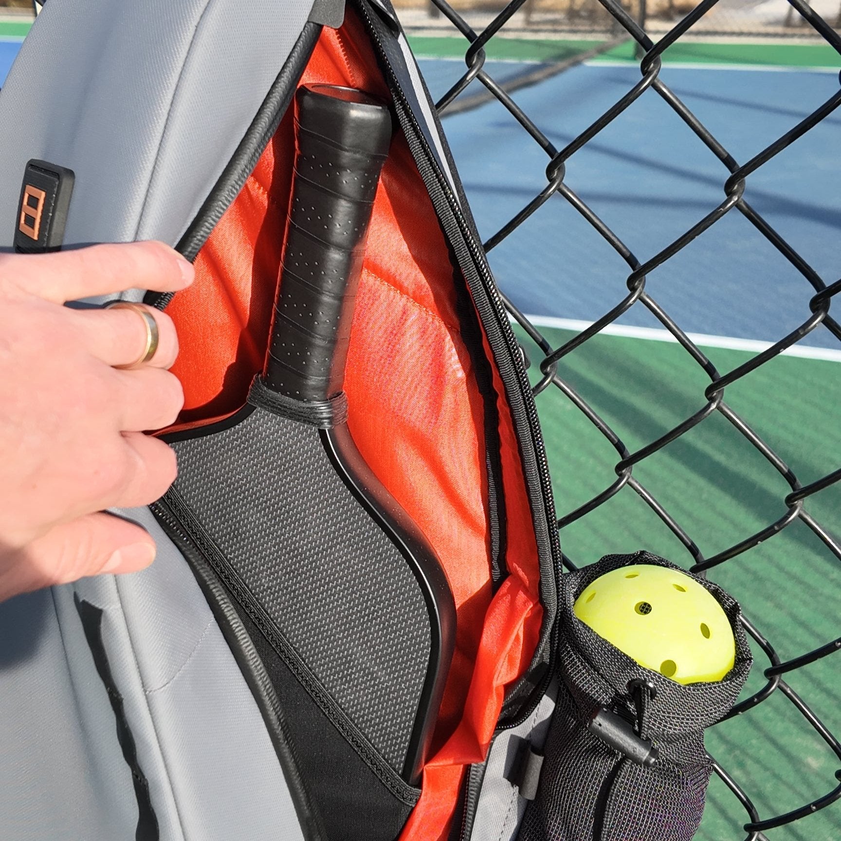 Pickleball racket in open gray and orange bag, with yellow ball in side pocket, next to chain-link fence. 
