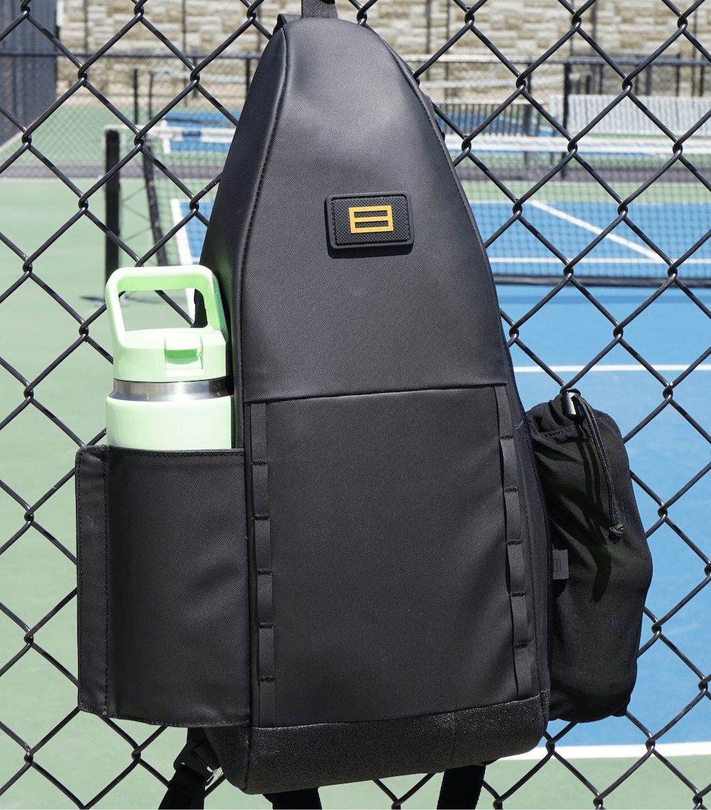 Black pickleball sling by FORWRD, with a yellow logo and side pockets holding a green water bottle and a black mesh pouch, hanging on a court fence.