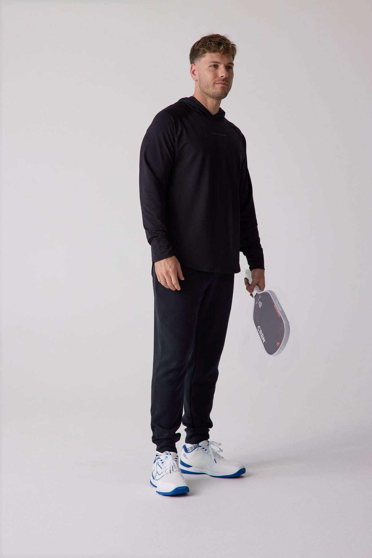 Black men's layering hoodie by Centerline, side view, with subtle logo, paired with matching joggers and white sneakers, holding paddle.