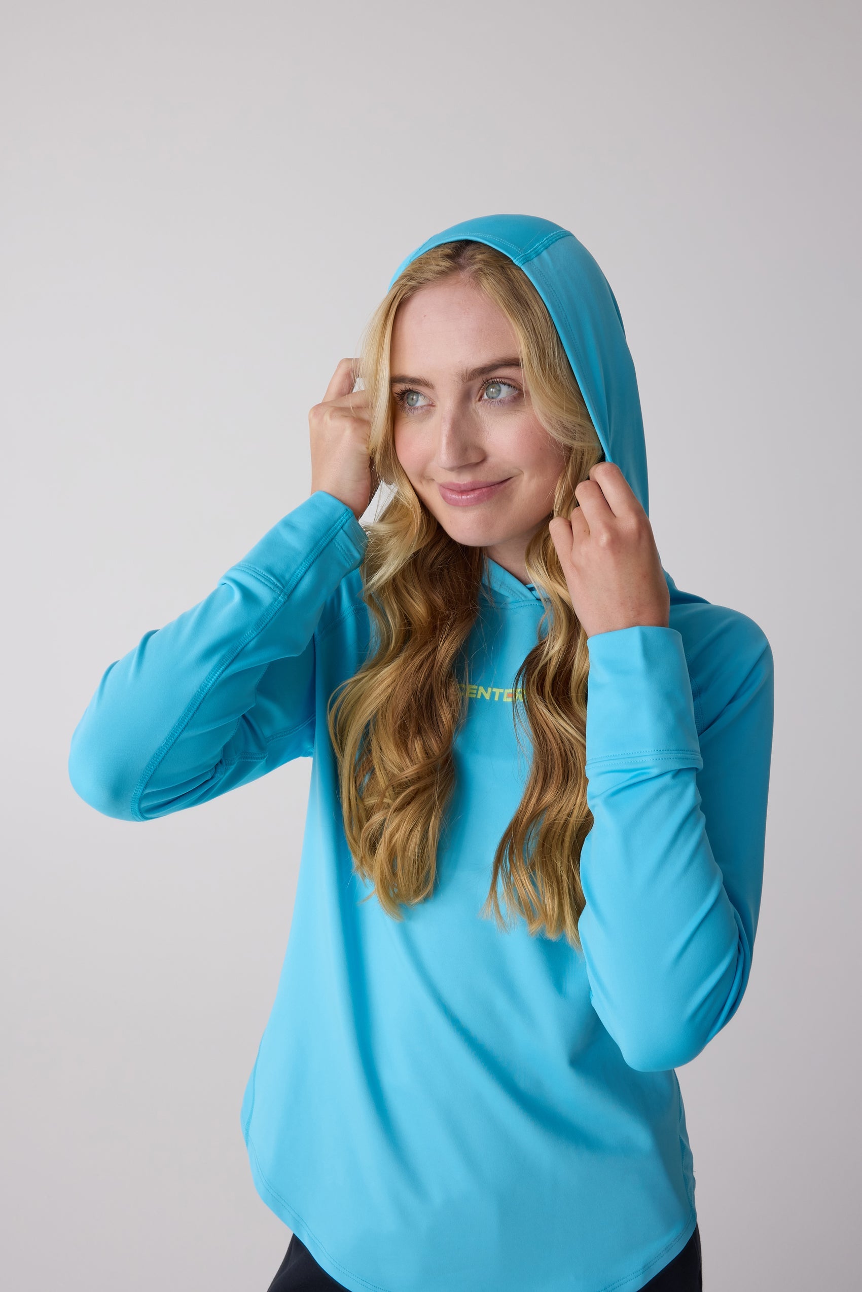 Blue smooth women's layering hoodie by Centerline Athletics, featuring yellow chest text, worn by a blonde woman adjusting the hood.