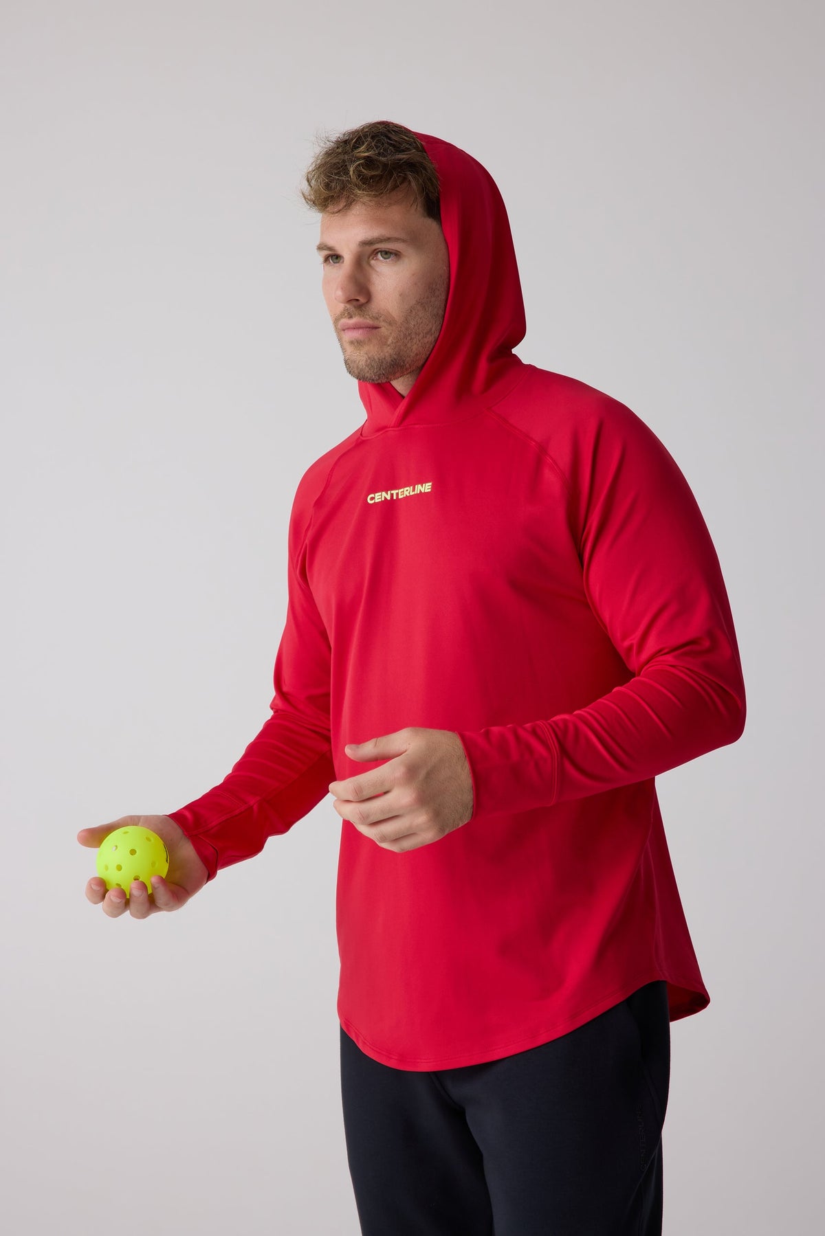Red men's layering hoodie by Centerline with white logo, side view, hood up, holding yellow ball
