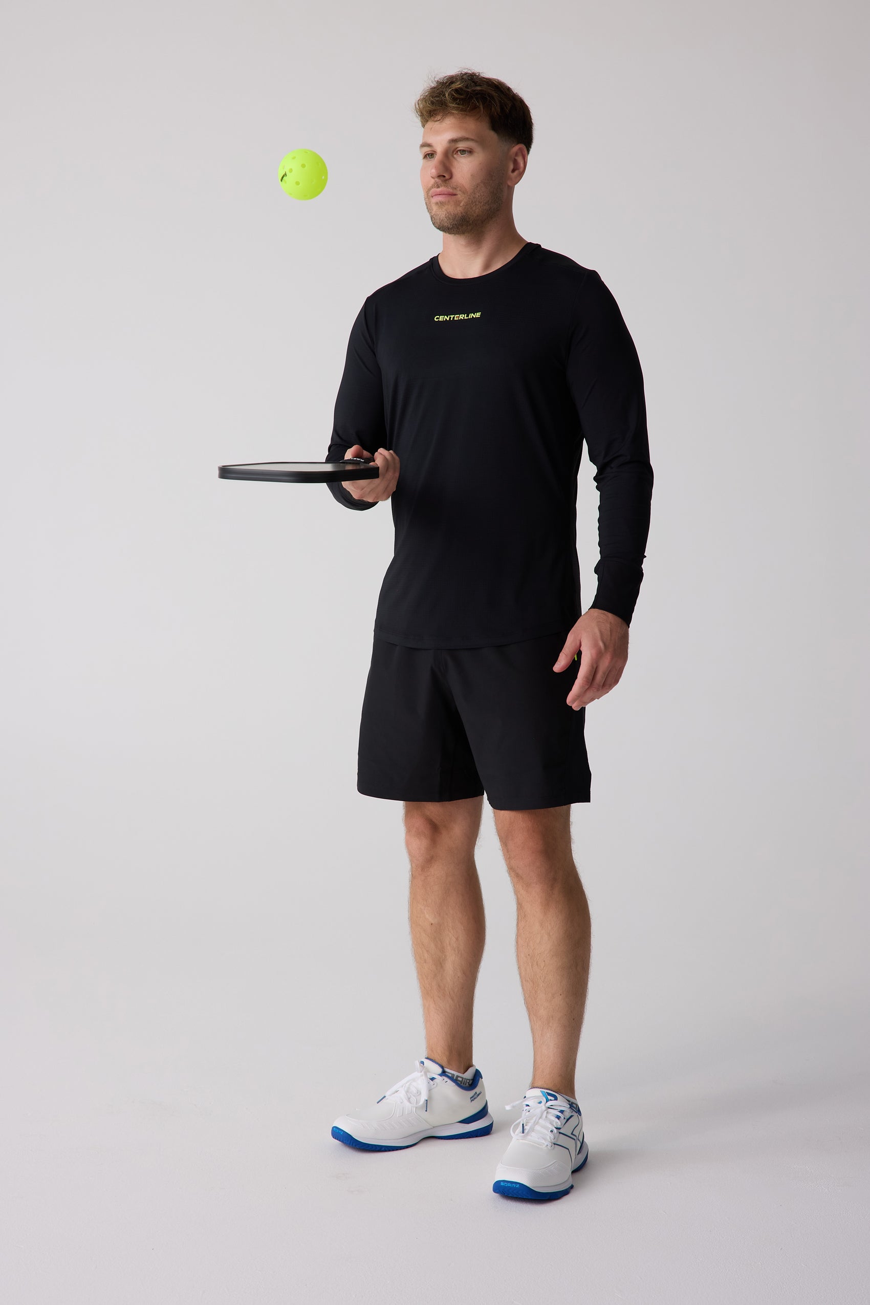 Black long sleeve shirt by Centerline Athletics with 'CENTERLINE' logo in neon green, worn by a man holding a paddle with a pickleball in the air.
