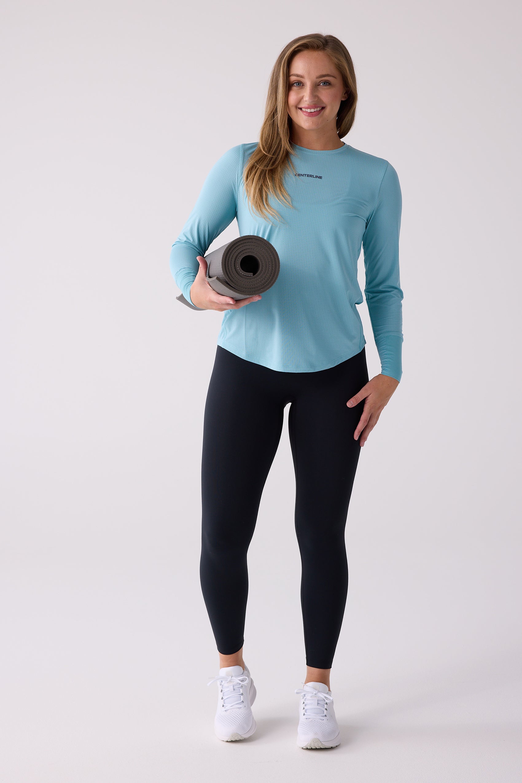 Light blue long sleeve shirt by Centerline Athletics with perforated fabric and chest logo, worn with black leggings, white shoes, and holding a yoga mat.